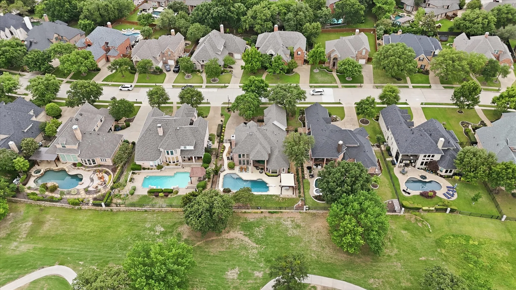 Aerial perspective showcasing a residential street lined with mature trees, featuring multiple properties with private swimming pools and extensive landscaping
