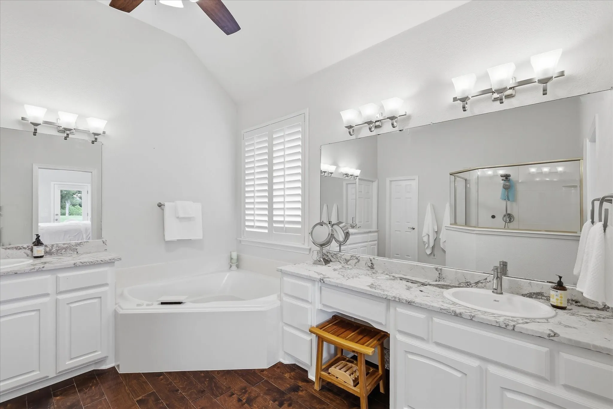 Spacious bathroom featuring a corner soaking tub, dual vanities with stone countertops, white cabinetry, and wood-finish flooring