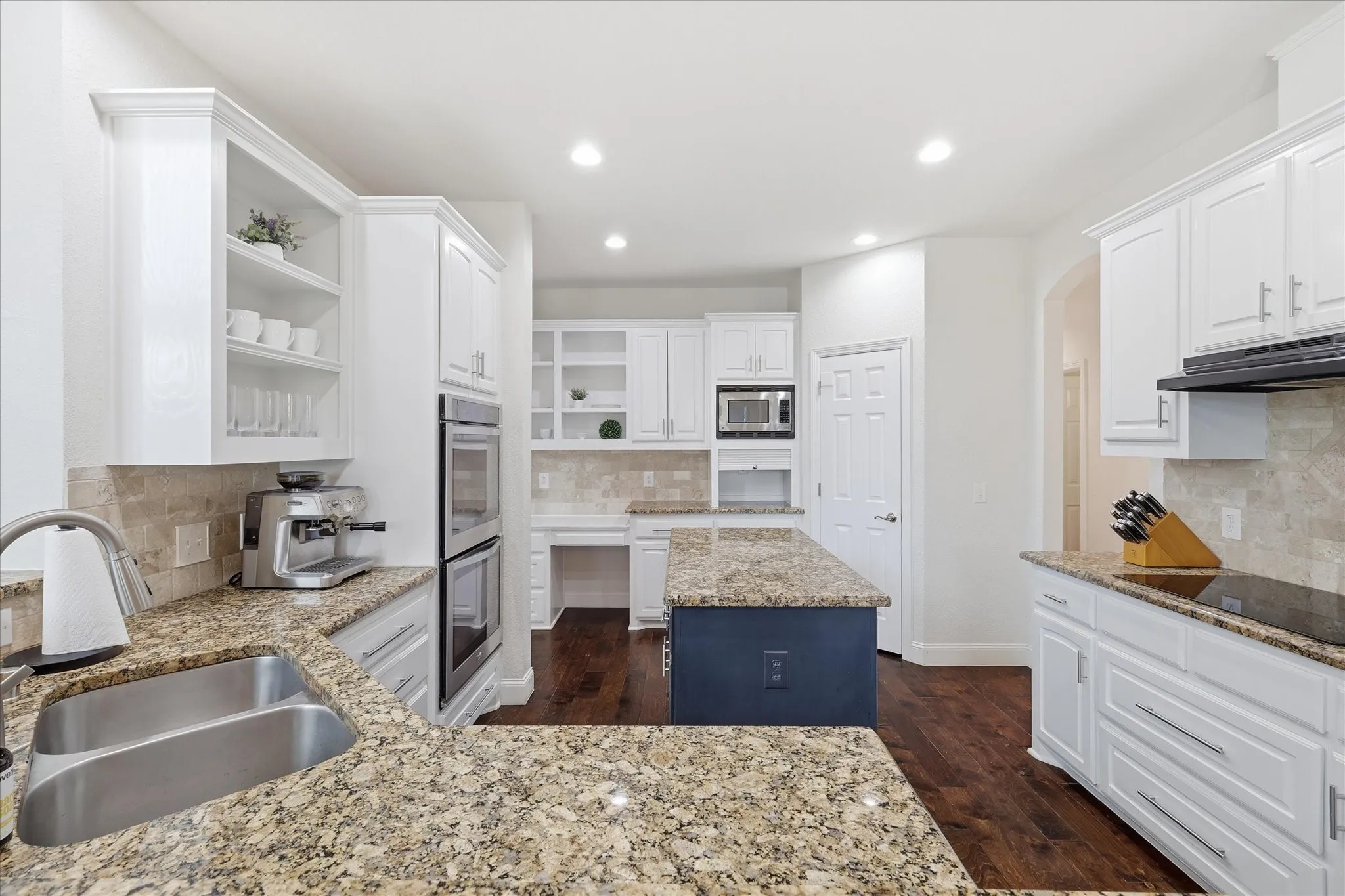 Kitchen featuring extensive light-toned granite countertops, white cabinetry with crown molding, double wall ovens, a center island with a contrasting base, and dark wood-finish flooring