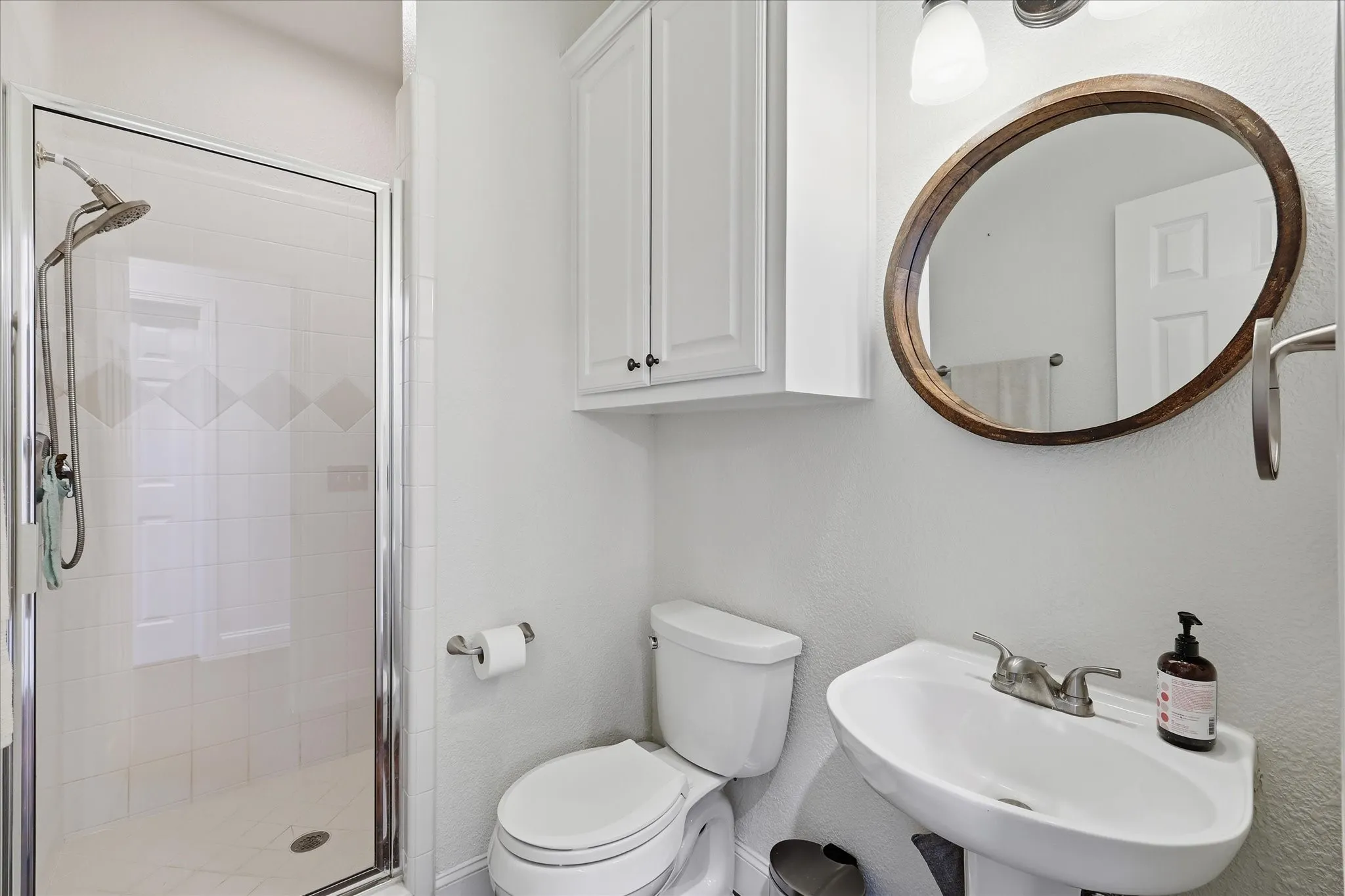 Bright bathroom featuring a glass-enclosed shower with tile surround, a pedestal sink with a two-handle faucet, a toilet, and an oval mirror with a wood-finish frame