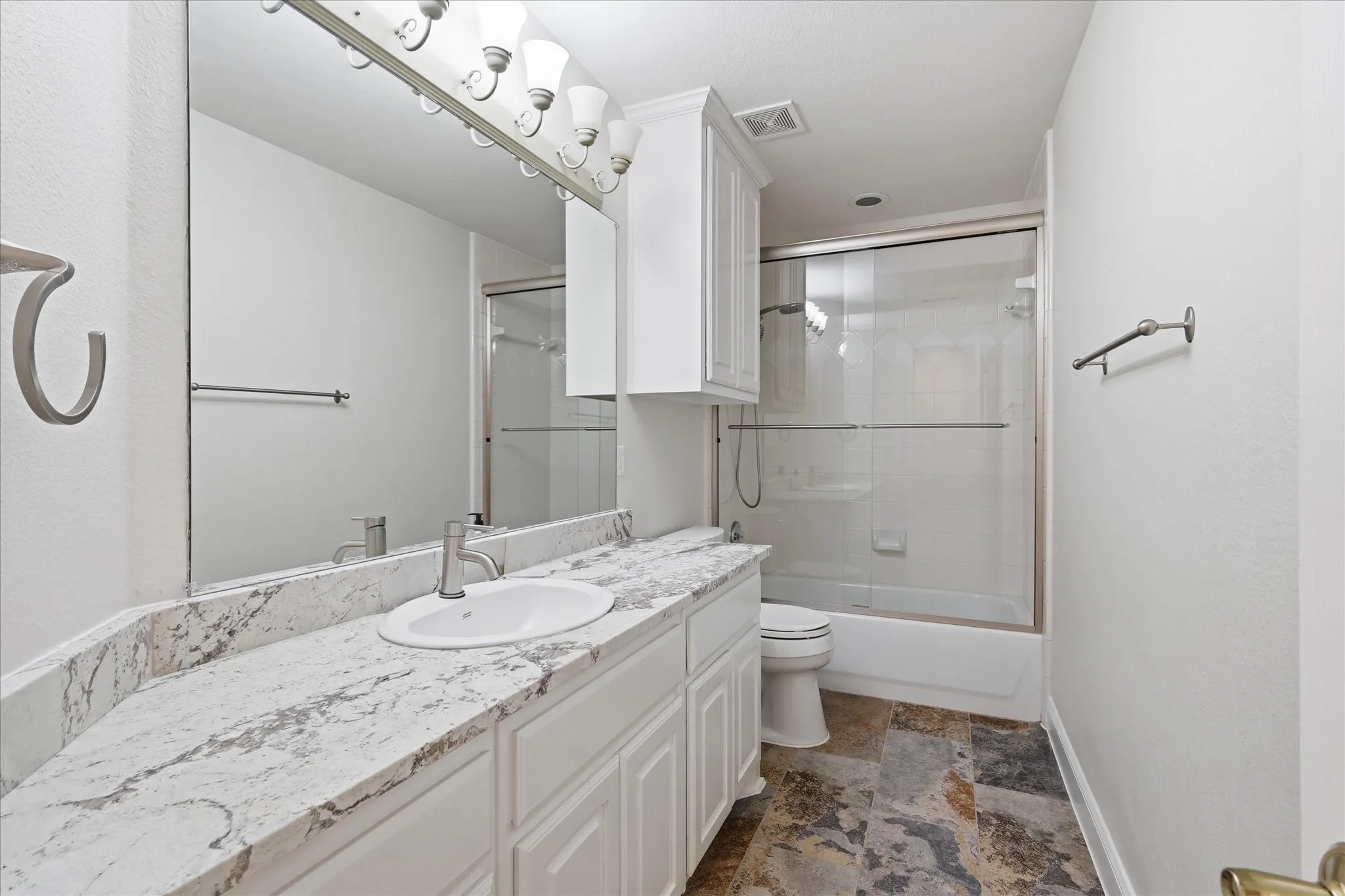 Bathroom featuring a vanity with a white countertop and undermount sink, white cabinetry, an overhead light fixture, a mirrored wall, and a shower-tub combination with a glass enclosure