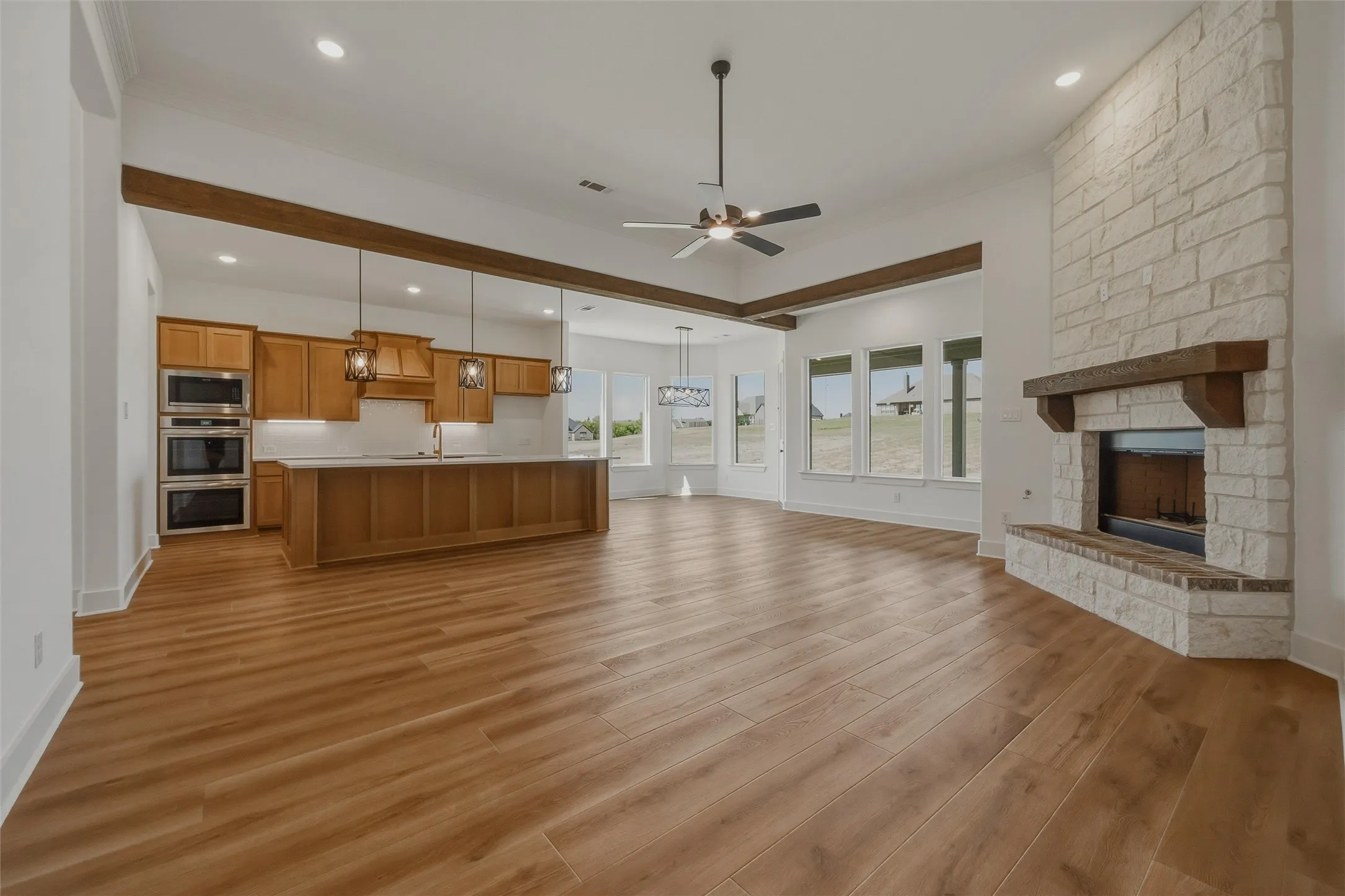 Open-concept living space featuring wood-finish flooring, a stone fireplace with a wood mantle, an island kitchen with a built-in oven, and a ceiling fan