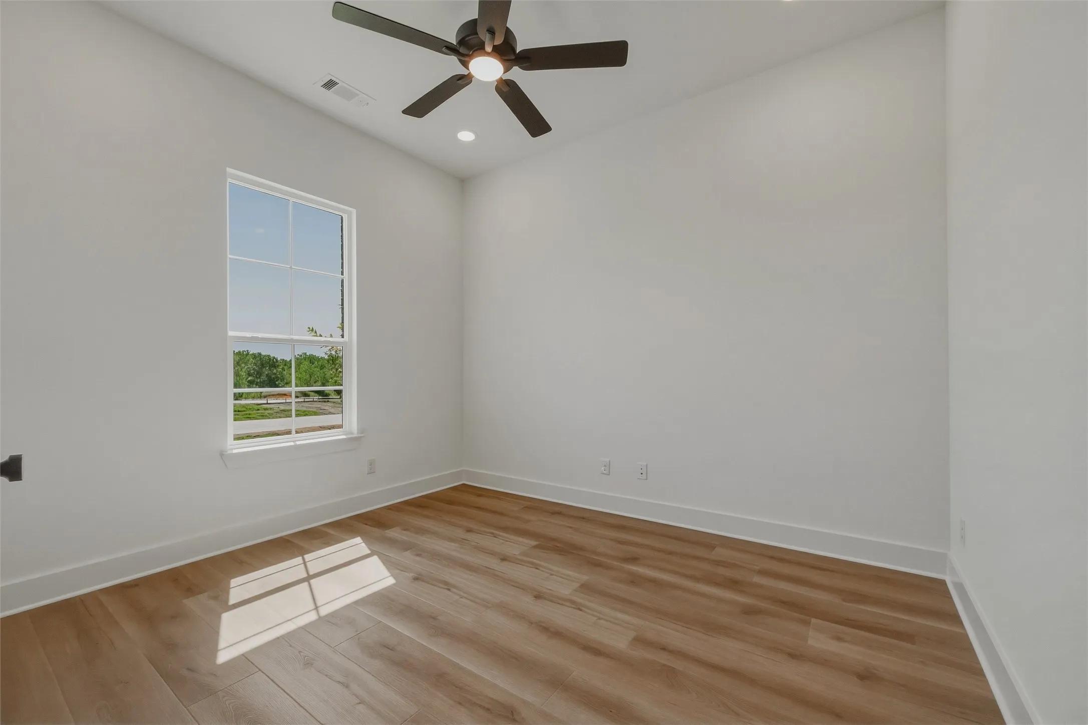 Spacious room with wood-finish flooring, a white-framed window, and a ceiling fan with integrated lighting