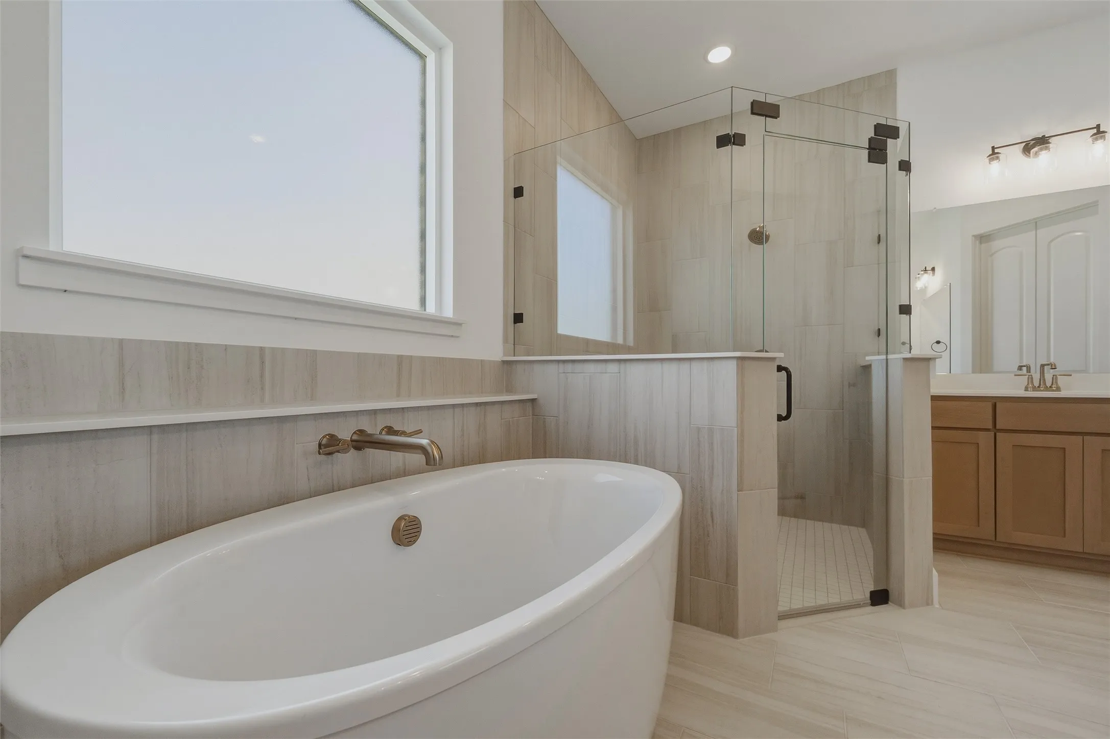 Freestanding soaking tub with wall-mounted faucet, glass-enclosed shower, tiled flooring, and dual vanity with integrated sinks