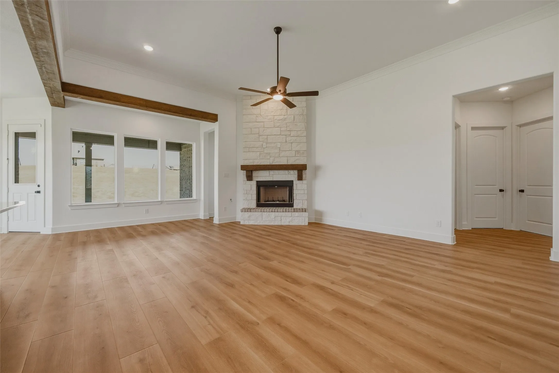 Vaulted ceiling with exposed wood beams, wood-finish flooring, stone-clad fireplace with wood mantel, ceiling fan, and multiple windows