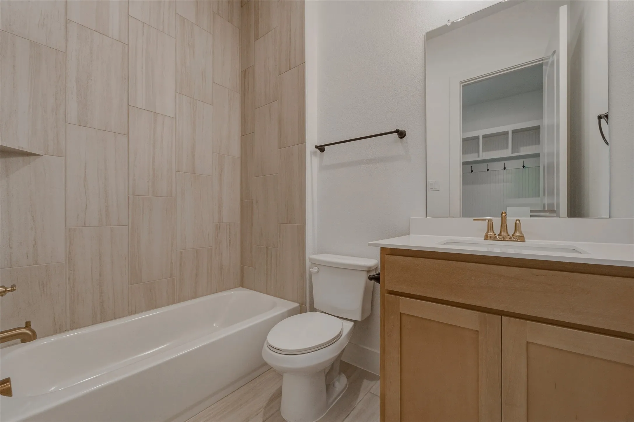 Full bathtub with a built-in tiled niche, wood-finish flooring, light-toned wood vanity, white countertop with an integrated sink, and a large wall-mounted mirror