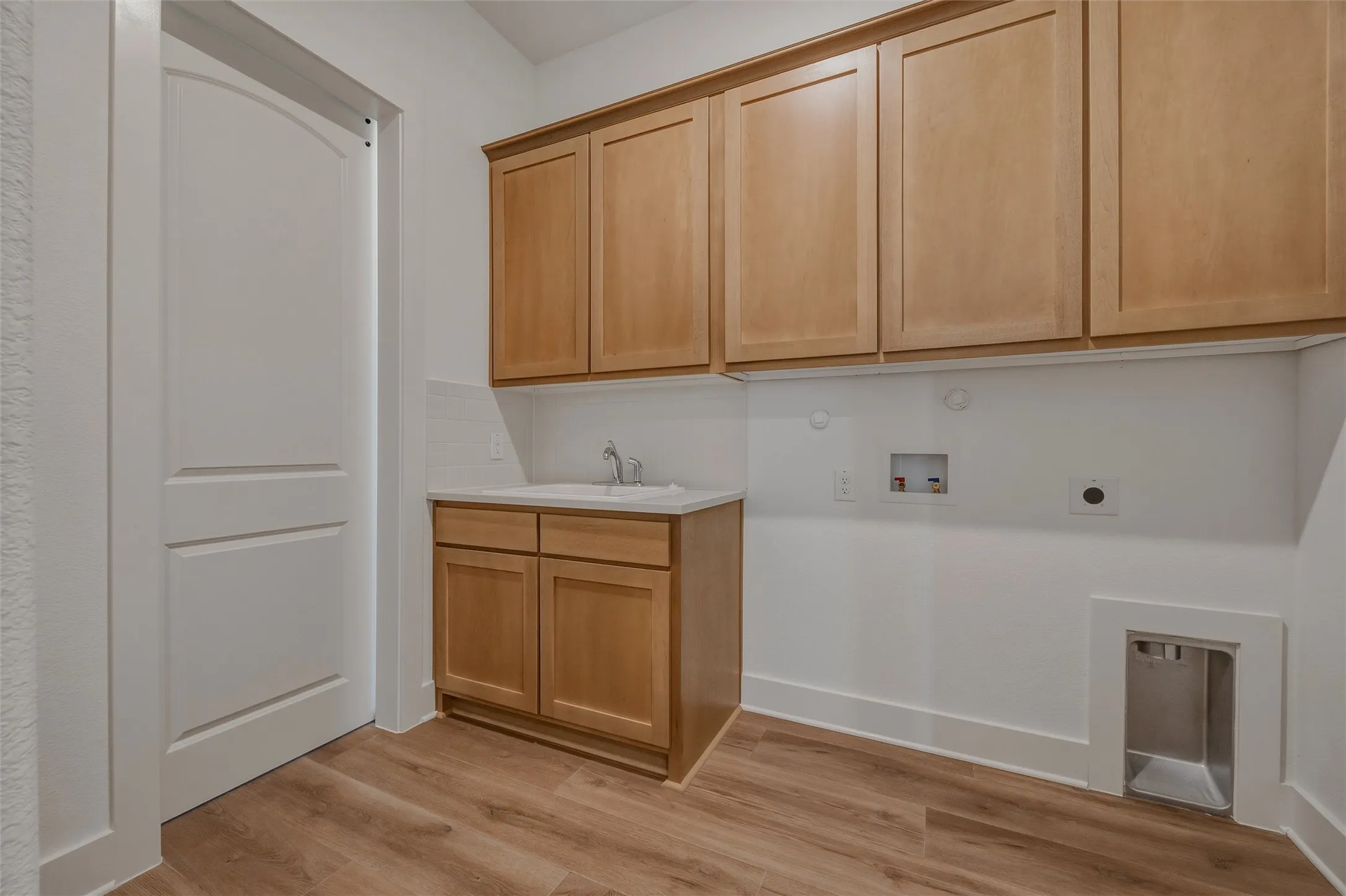 Wood-finish flooring, light wood cabinetry, white countertop with integrated sink, white tiled backsplash, and white interior door