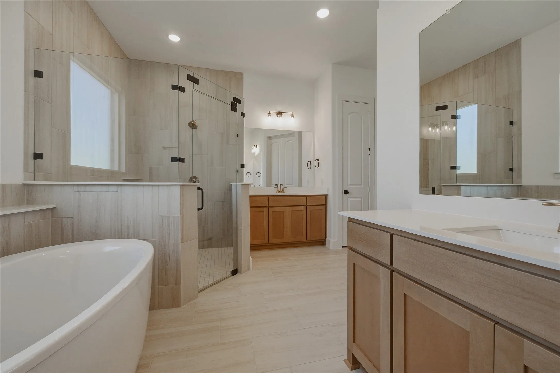 Freestanding soaking tub, glass-enclosed shower with tiled walls, dual vanities with integrated sinks, and light wood-finish cabinetry