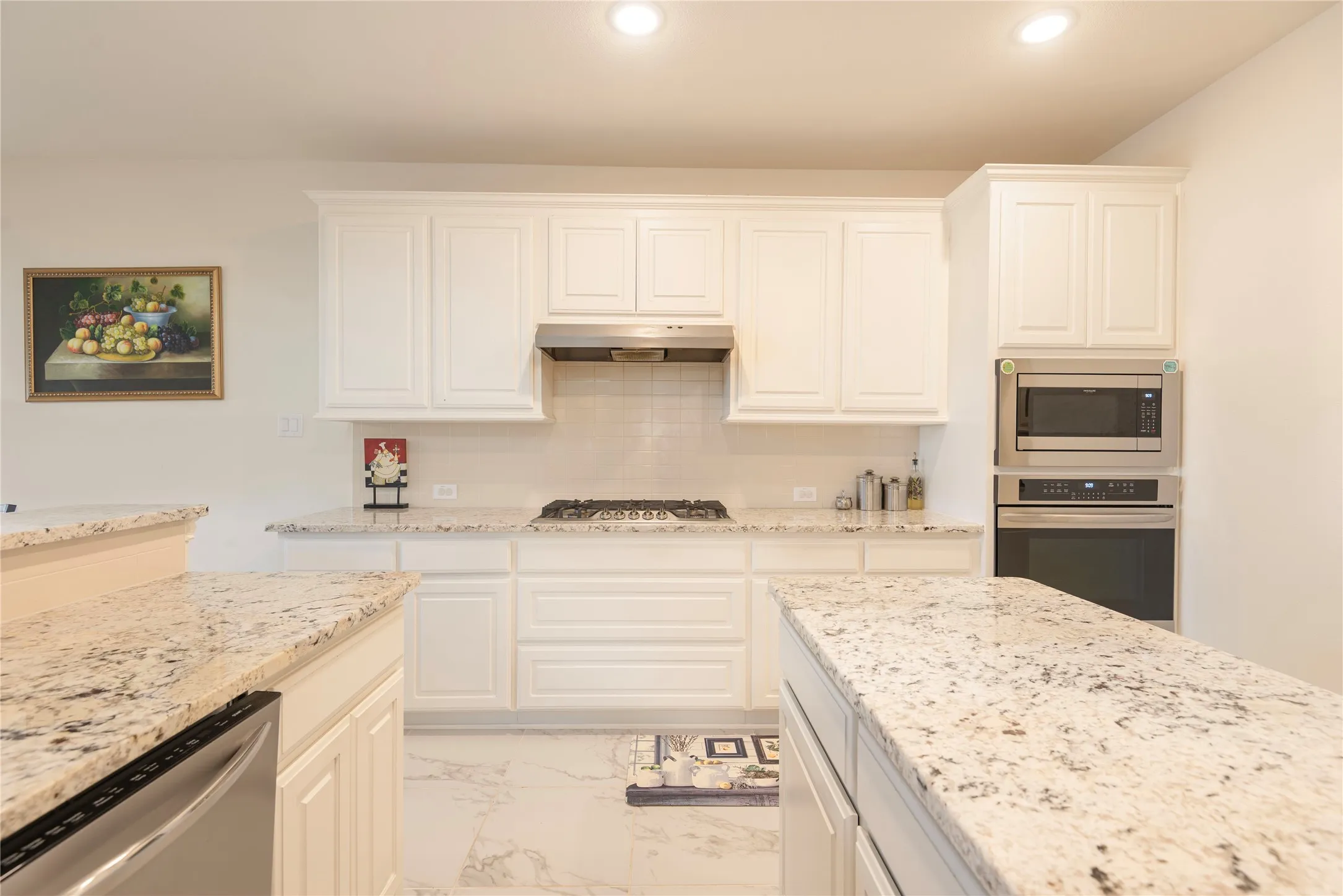 Modern kitchen featuring white shaker-style cabinetry, light-toned stone countertops, and stainless steel appliances including a built-in oven and microwave