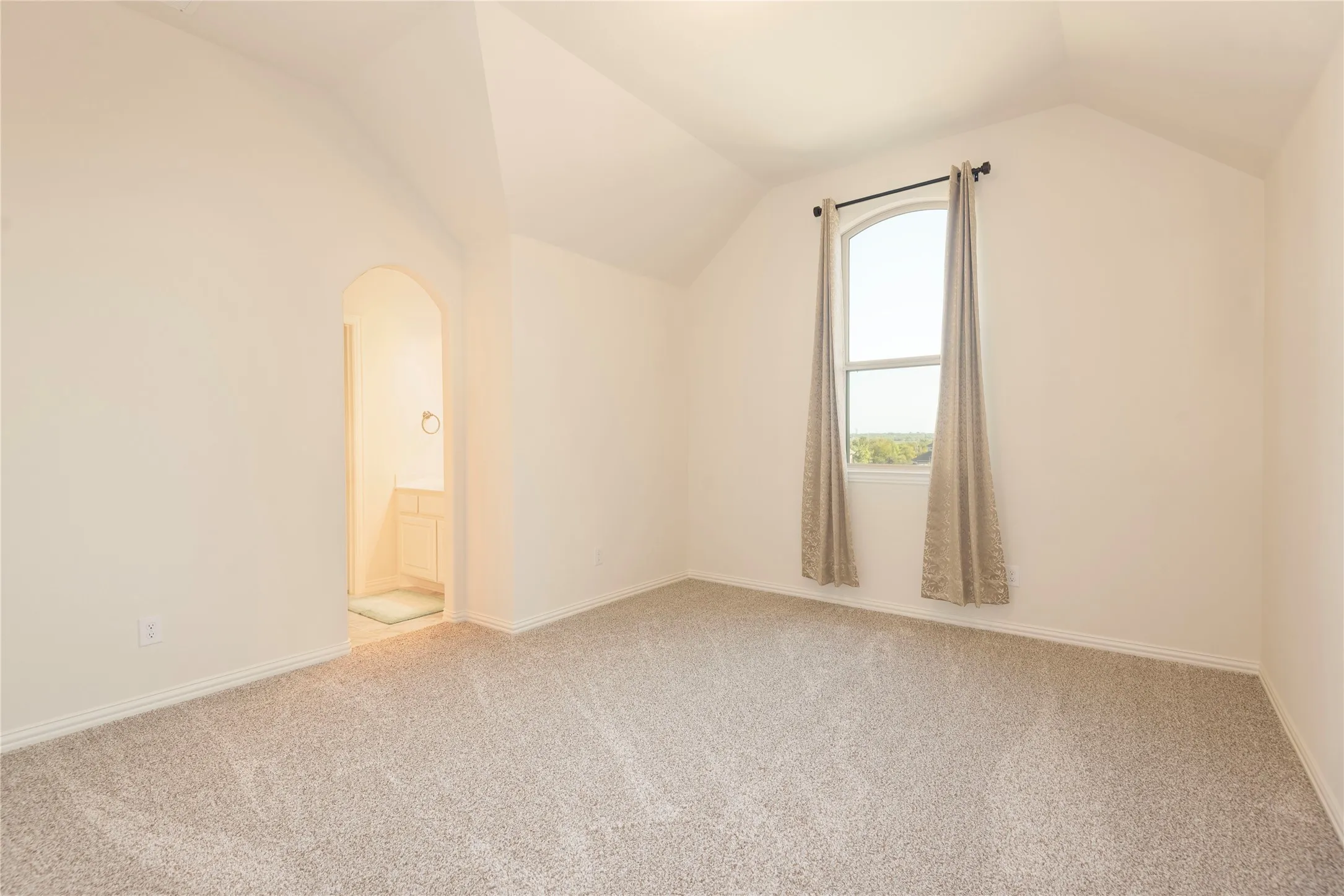 Spacious room featuring vaulted ceilings, light-colored carpeting, and an arched window with neutral drapery