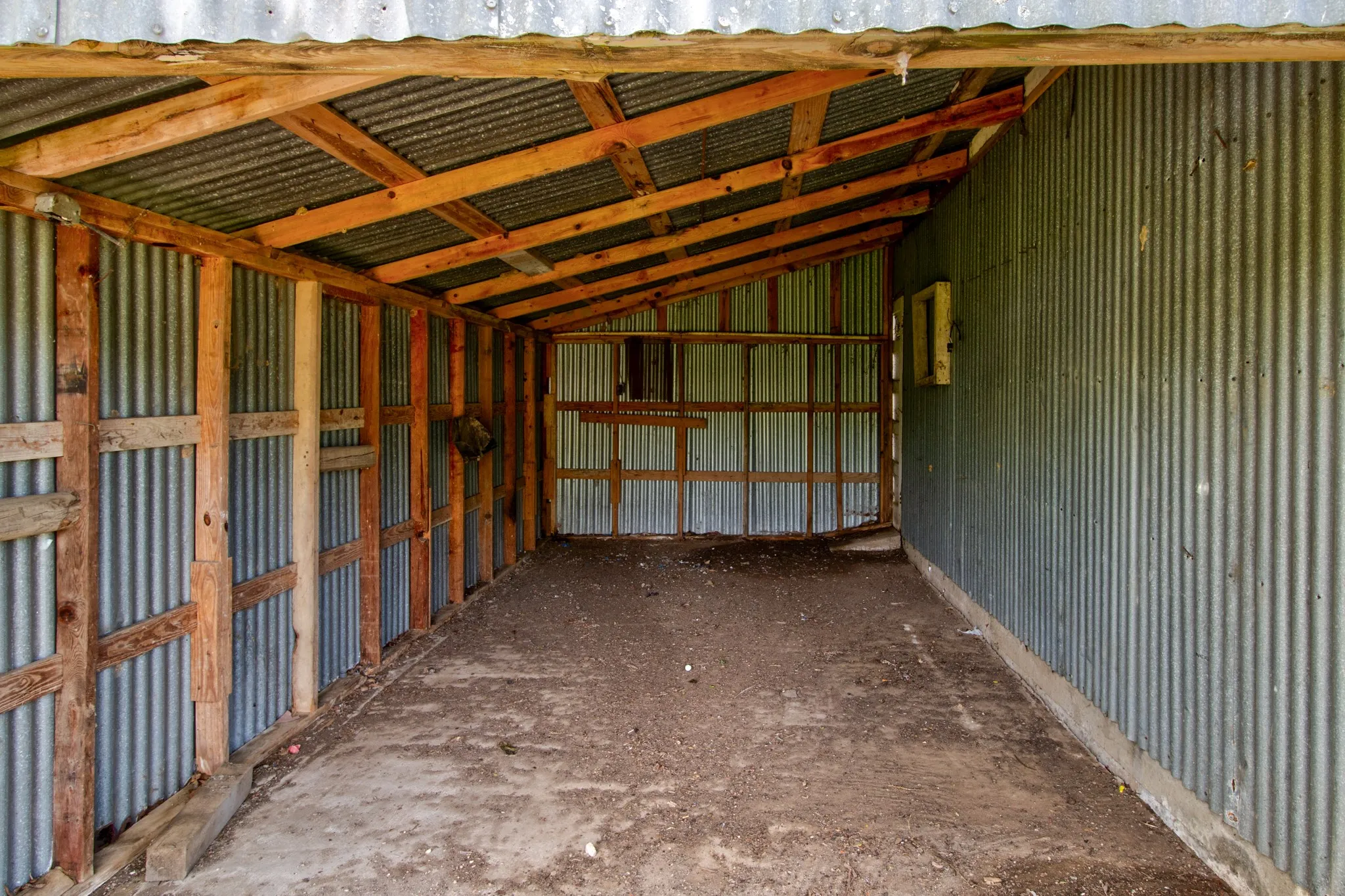 Open-sided shed structure featuring a corrugated metal roof and walls, timber framing, and an earthen floor