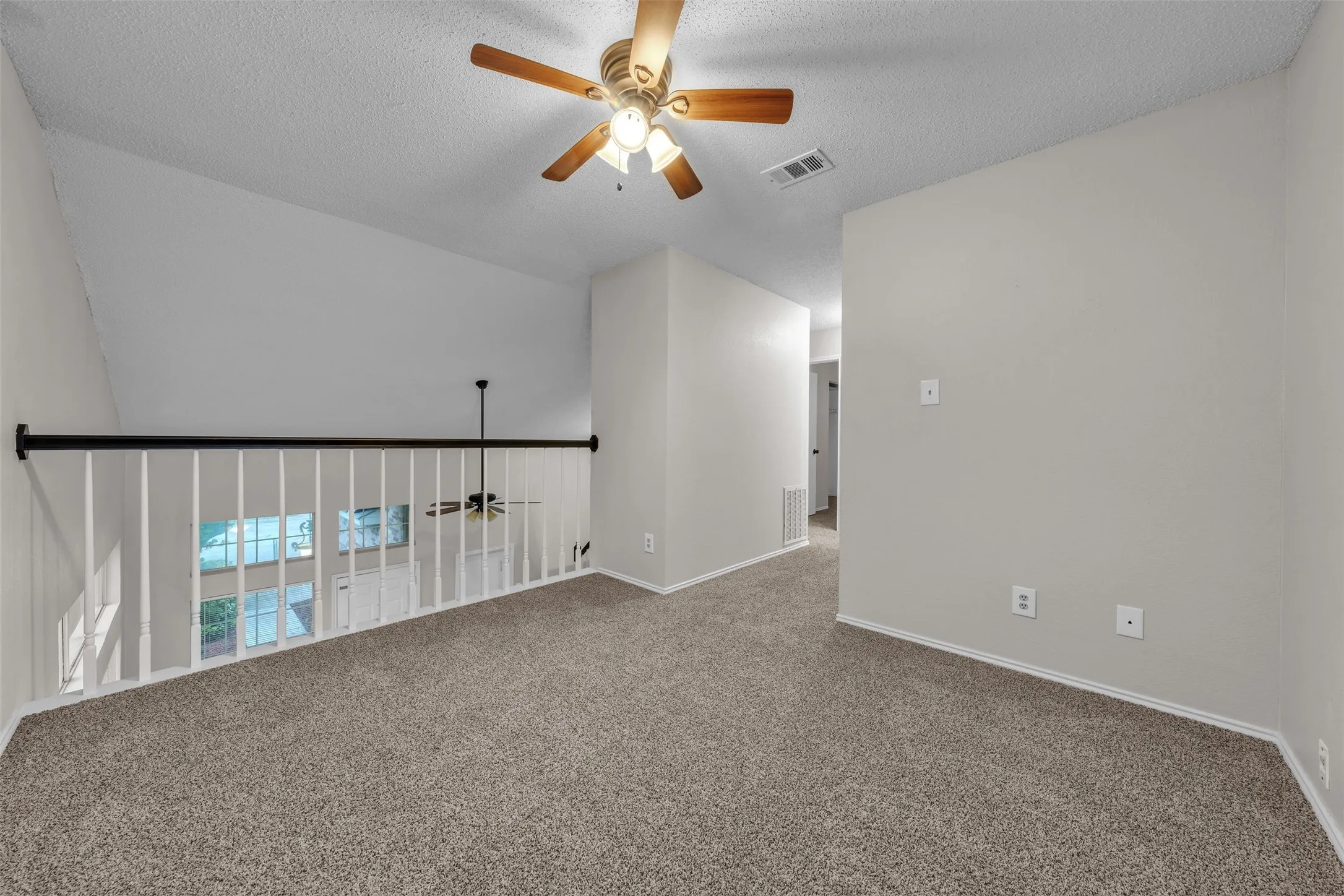Carpeted loft area featuring a white spindle railing with a black handrail, an angled ceiling line, a ceiling fan with integrated lighting, and multiple windows below