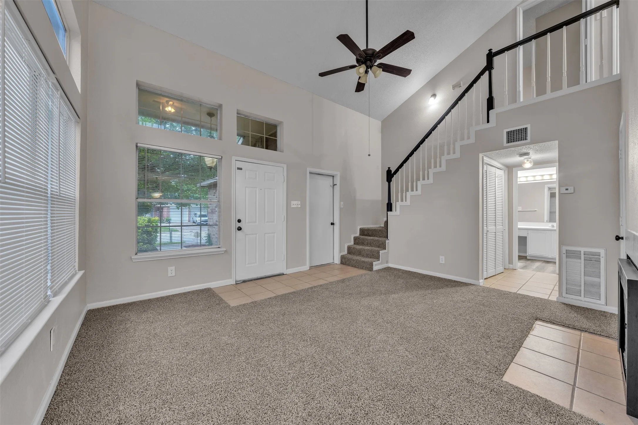 Spacious living area featuring high vaulted ceilings, expansive window array, a ceiling fan with light fixture, and a carpeted staircase leading to an upper level