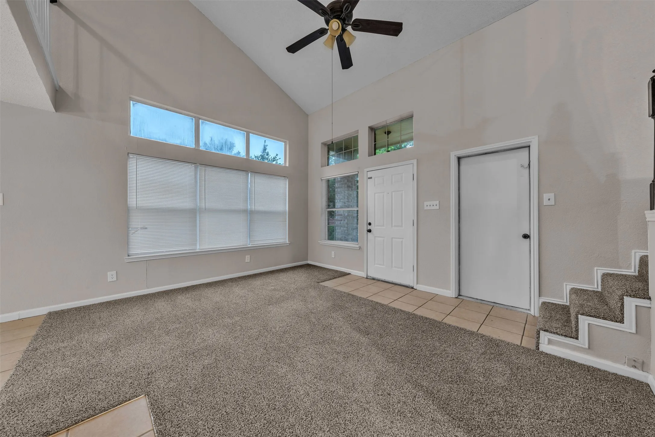 Vaulted ceiling great room featuring a ceiling fan, a wall of windows with upper transom windows, and carpet flooring