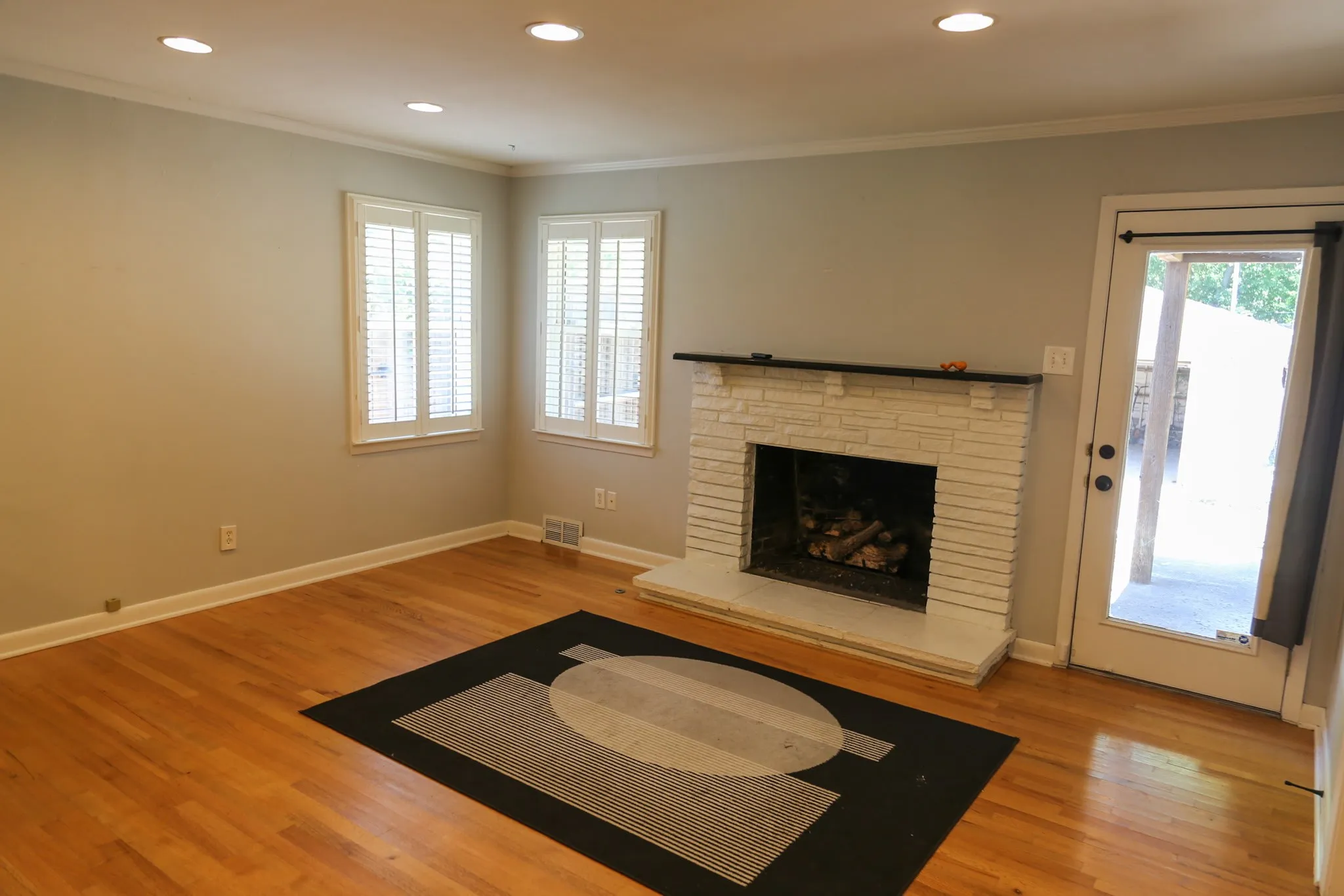 Living area featuring wood-finish flooring, a white brick fireplace with a black mantel, recessed lighting, and white plantation shutters on two windows