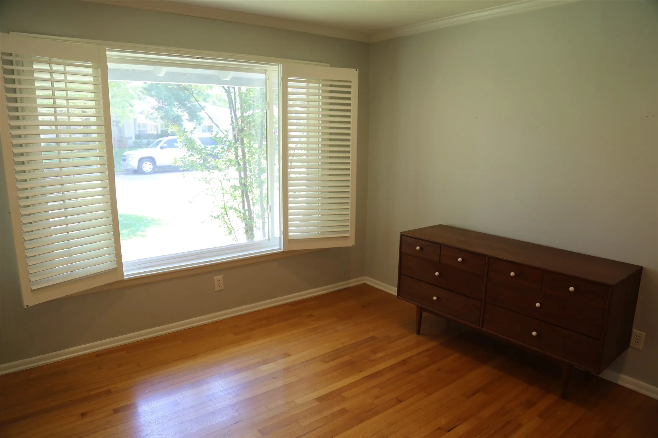 Hardwood flooring complemented by a prominent window featuring white plantation shutters and crown molding