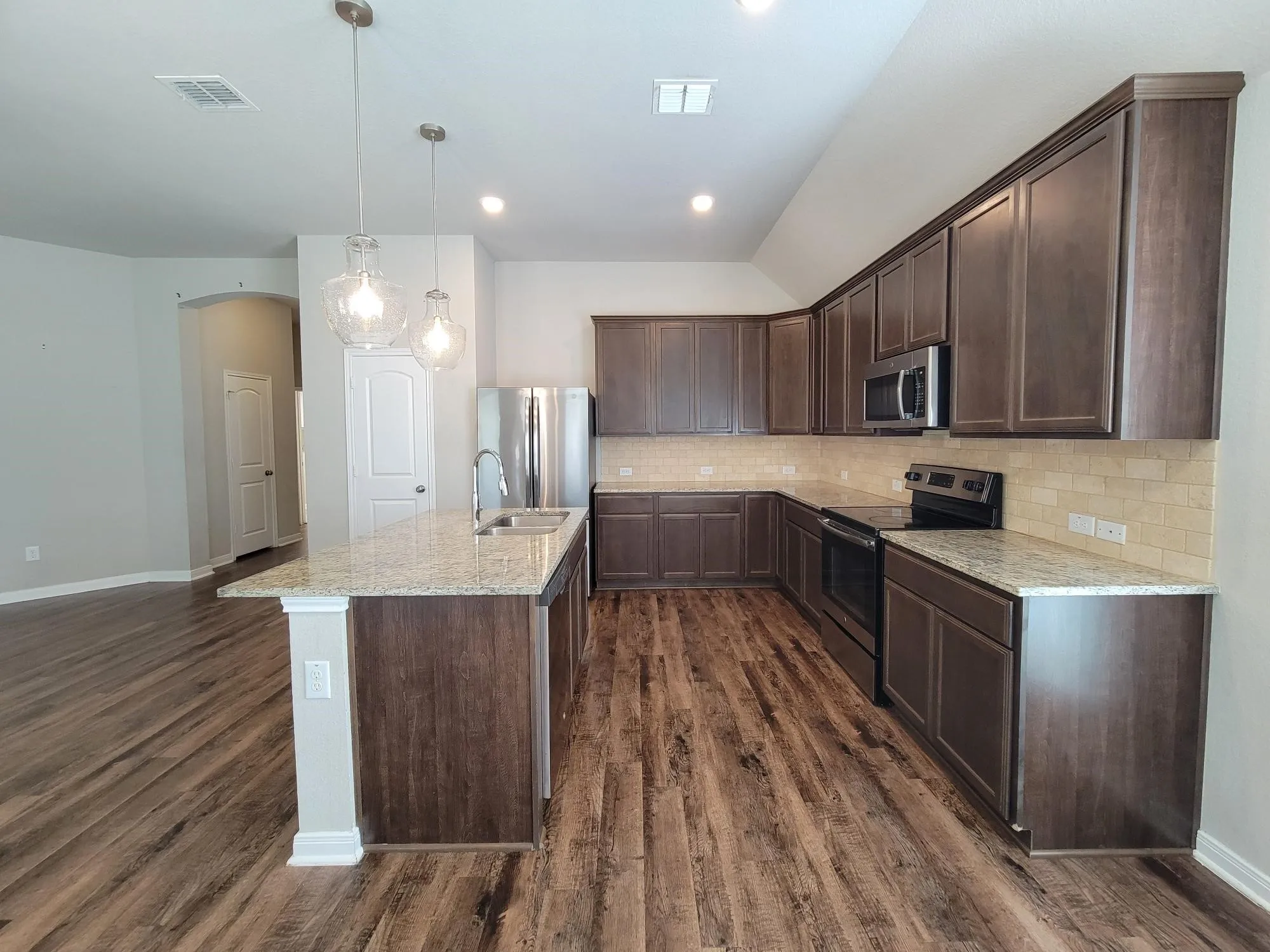 Open-concept kitchen featuring wood-finish flooring, a central island with a sink, pendant lighting, shaker-style cabinetry, and stone countertops