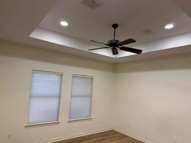 Tray ceiling with recessed lighting, dark wood-finish ceiling fan, two windows with blinds, and wood-finish flooring