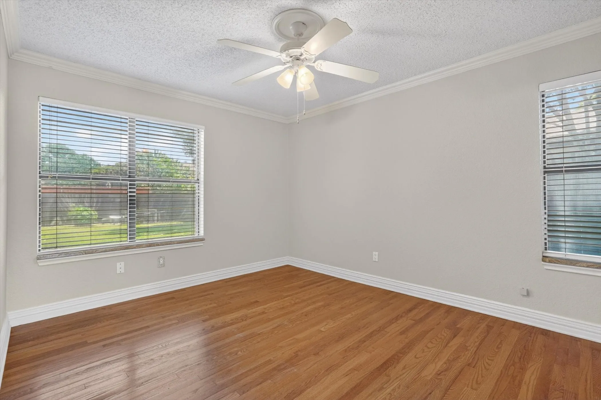 Spacious room featuring wood-finish flooring, two windows with blinds, a ceiling fan with integrated lighting, and crown molding