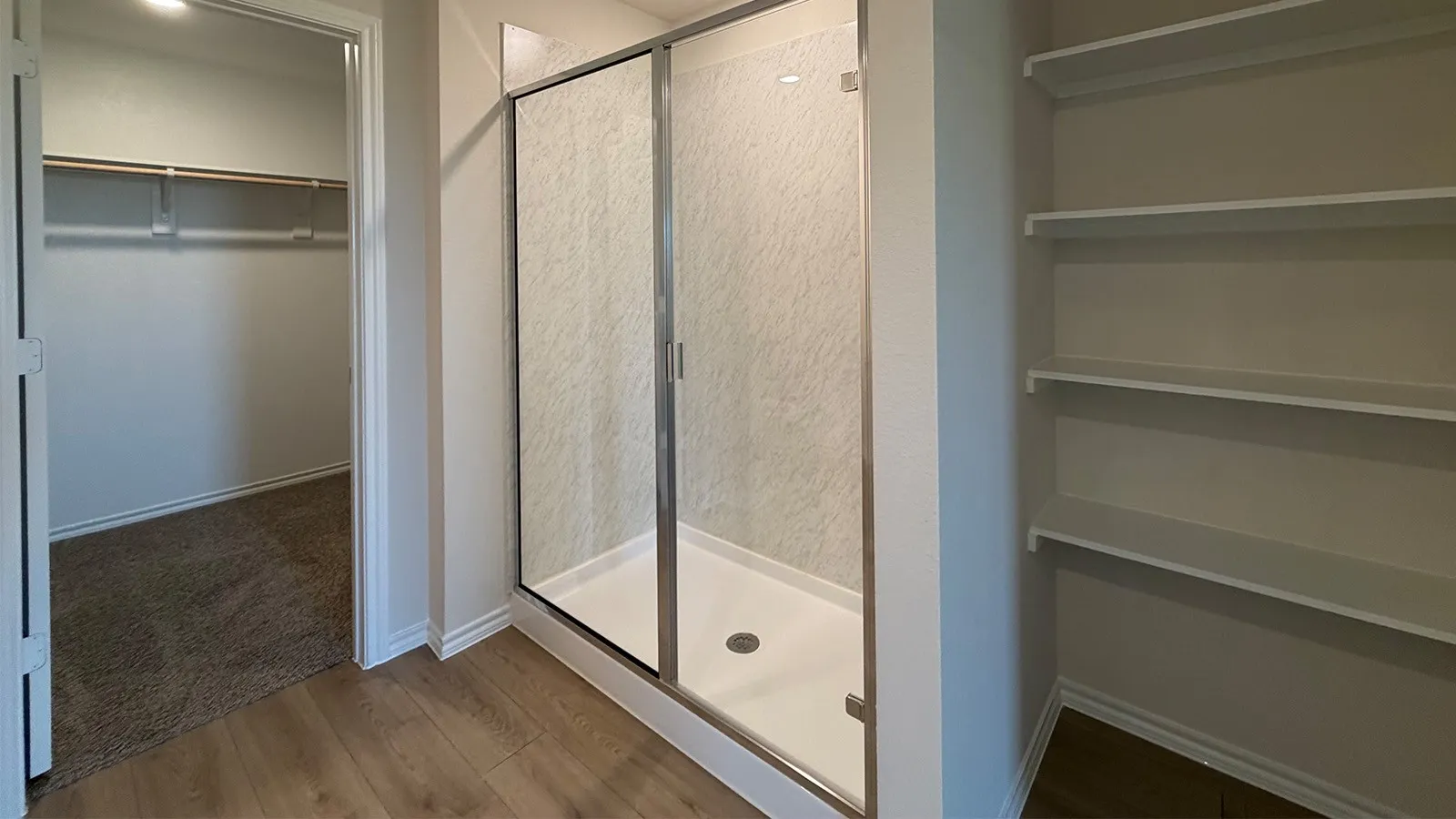 Walk-in shower with clear glass enclosure and textured wall panels, wood-finish flooring, built-in shelving, and an additional walk-in closet with carpeted flooring and hanging rods