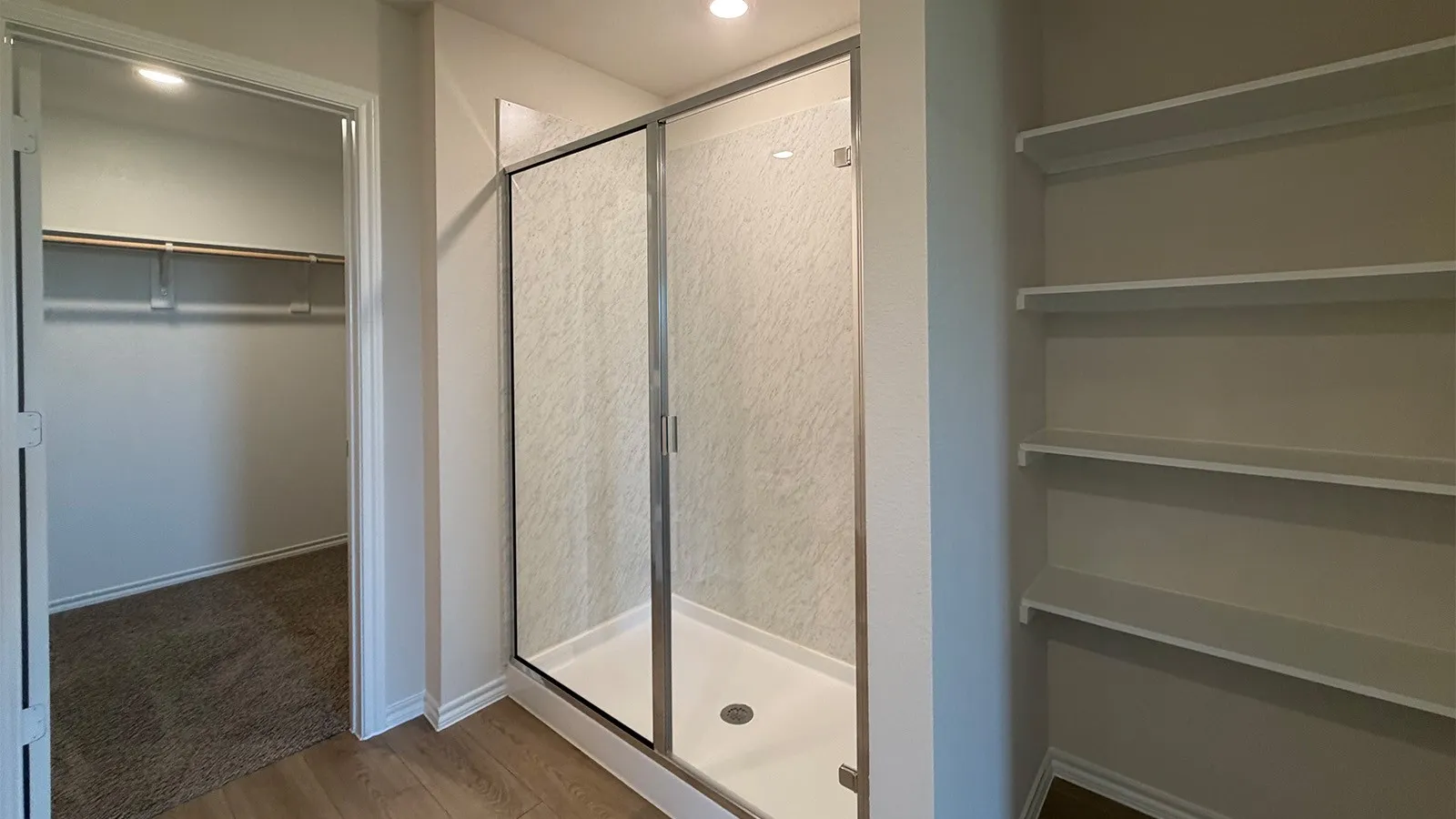 Glass-enclosed shower with a textured surround and white base, featuring wood-finish flooring, built-in shelving, and a separate walk-in closet with hanging rods