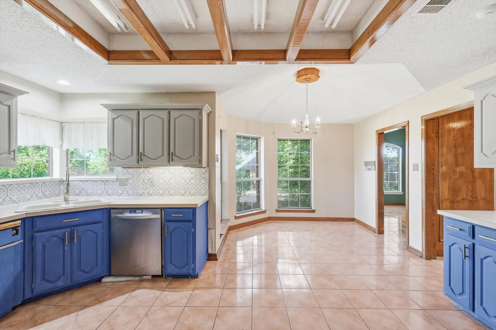 Kitchen and dining area featuring vibrant blue and grey cabinetry, stainless steel dishwasher, tile flooring, and a decorative ceiling with exposed wood beams