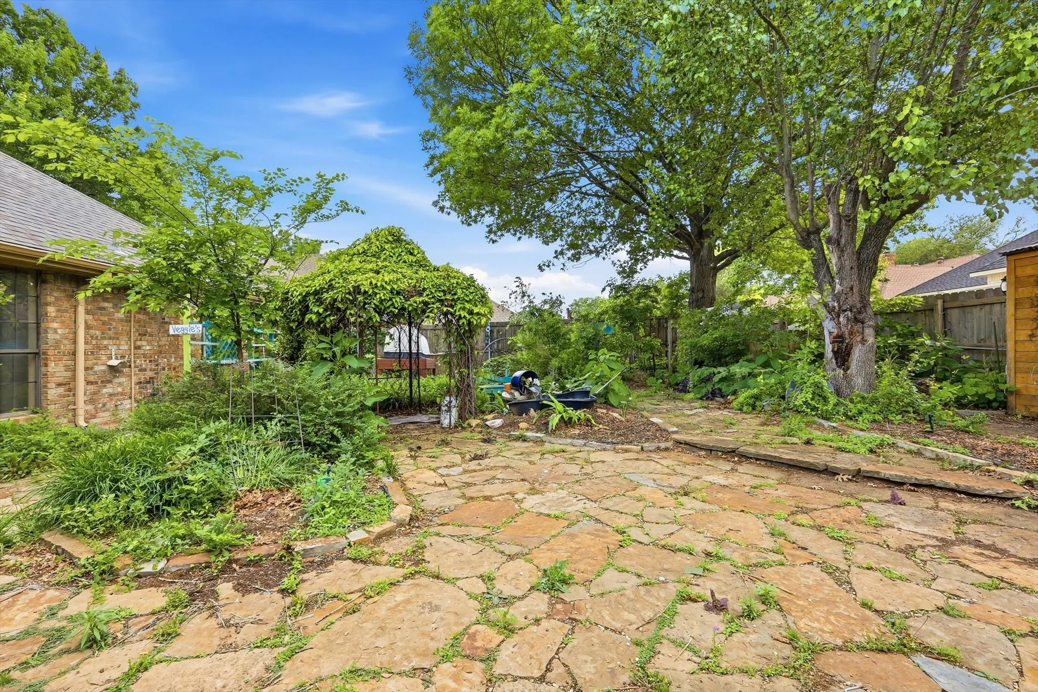 Expansive flagstone patio area featuring mature trees, a lush green vine-covered pergola, and a brick-sided exterior with a sloped roofline