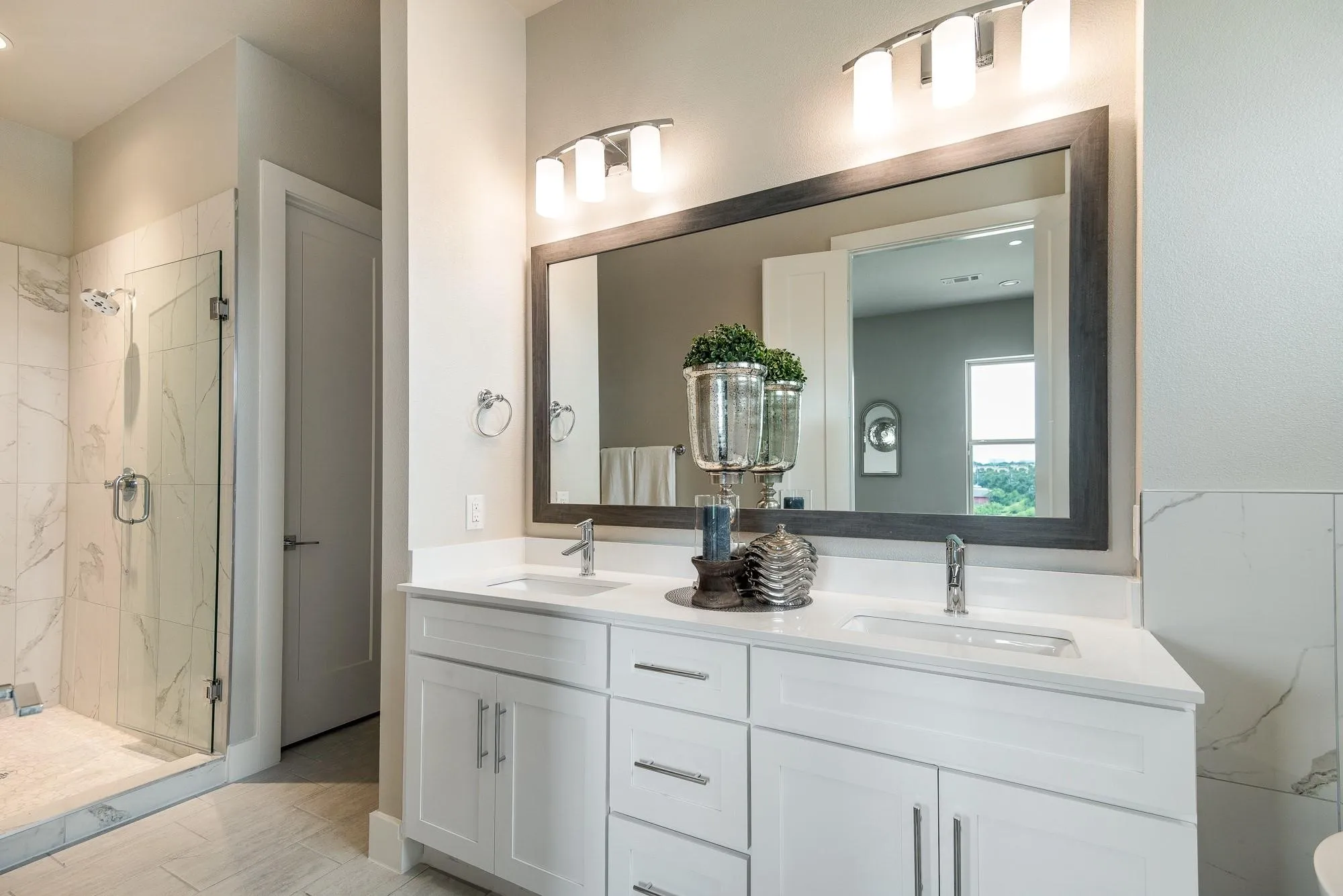 Well-appointed bathroom featuring a double vanity with white cabinetry and chrome fixtures, a large framed mirror, and a spacious walk-in shower with glass enclosure and marble-look tile