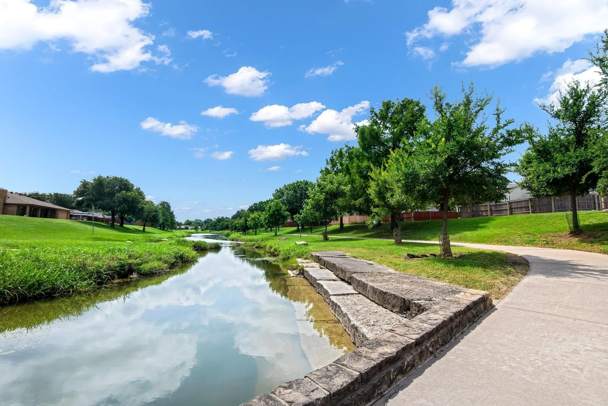 Scenic view of a waterway bordered by lush green grass, mature trees, and a paved pathway