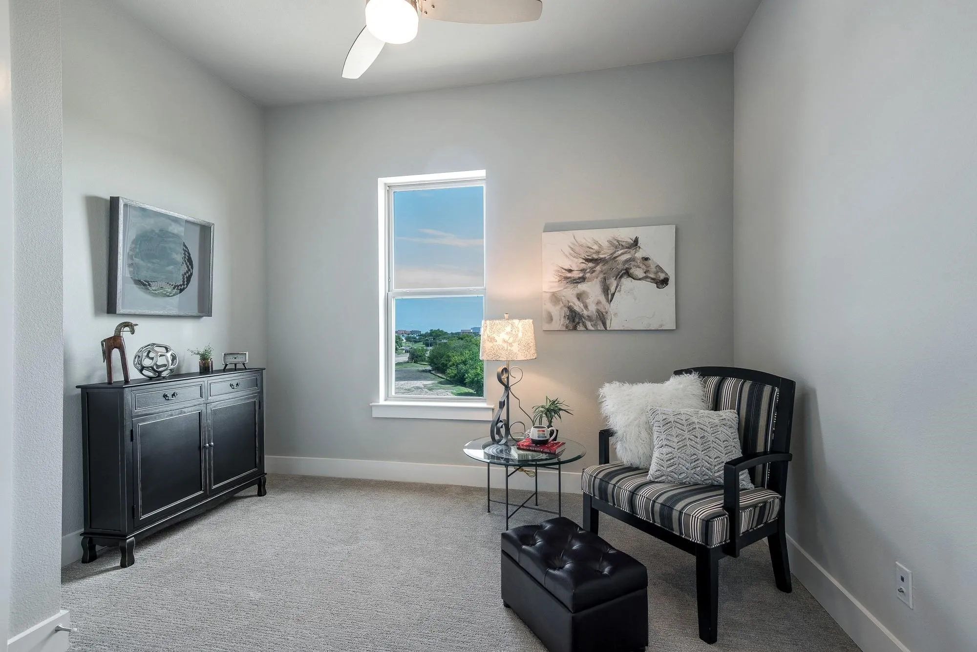 This room features light grey walls and light grey carpet, a window with a view of greenery and sky, and a ceiling fan