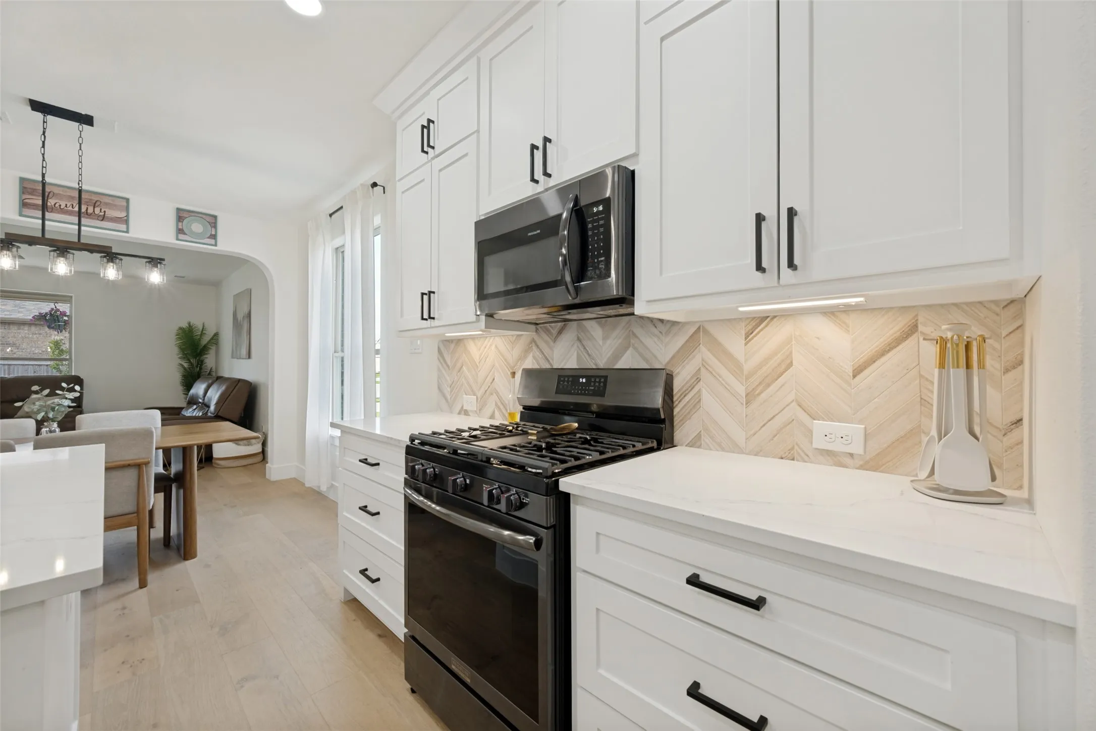 Kitchen featuring white shaker cabinetry with dark hardware, stainless steel microwave, gas range, wood-finish herringbone backsplash, and light-tone wood-finish flooring