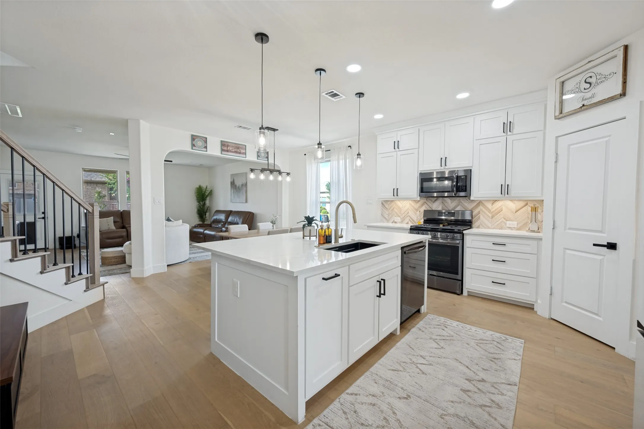 Open-concept kitchen featuring an island with an undermount sink, white cabinetry with dark hardware, stainless steel appliances, and wood-finish flooring