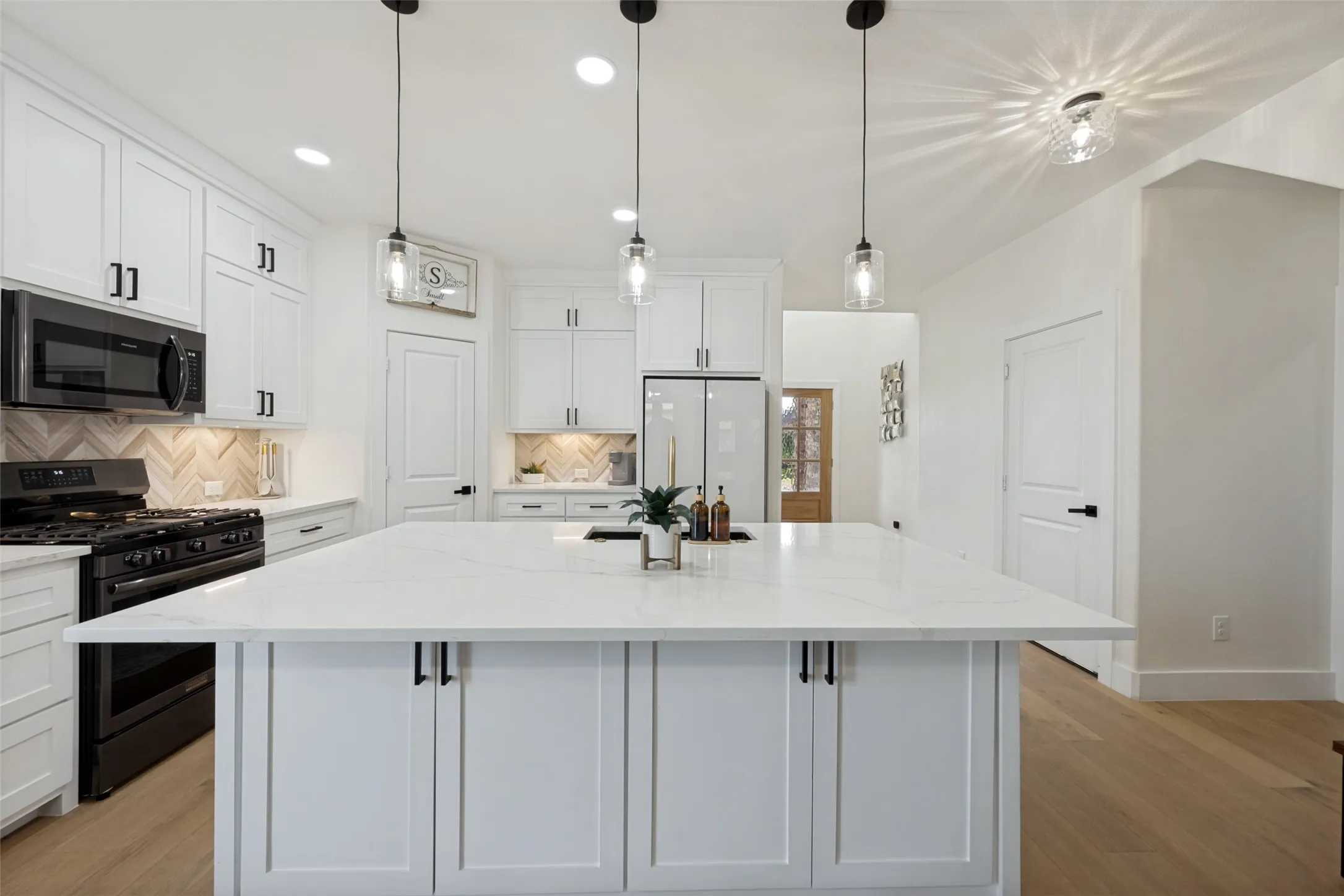 Kitchen featuring a large central island with a white countertop, white shaker-style cabinetry with black hardware, black appliances, a herringbone tile backsplash, and wood-finish flooring