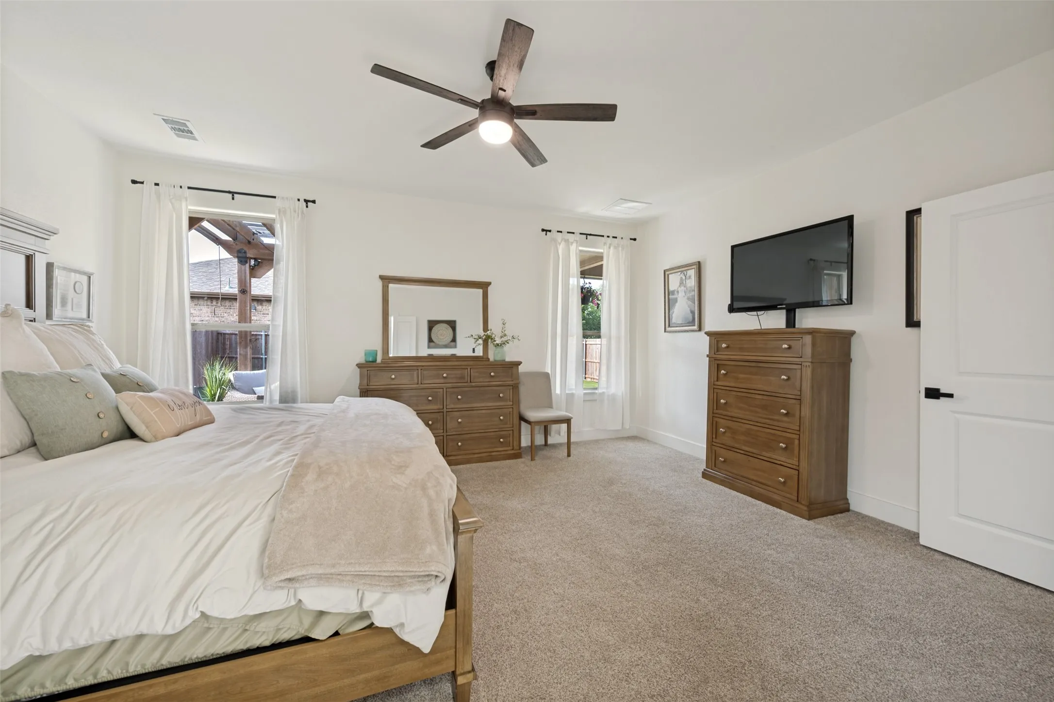 Spacious bedroom featuring light neutral wall paint, plush carpet flooring, a ceiling fan with integrated lighting, and two windows with white curtains providing natural illumination