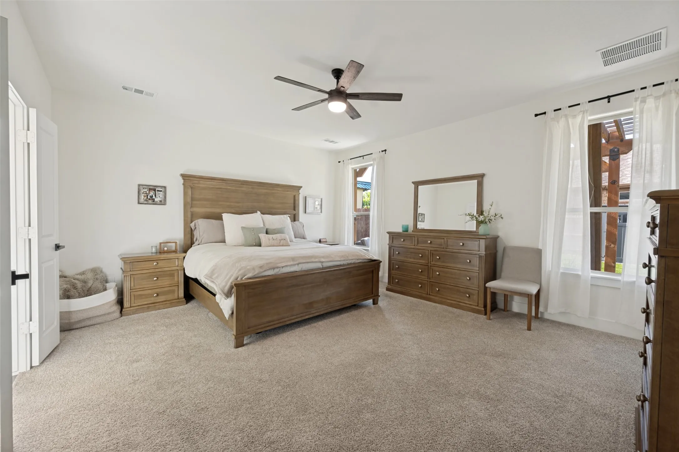 Carpeted primary bedroom featuring a ceiling fan, white walls, and two windows with white curtains