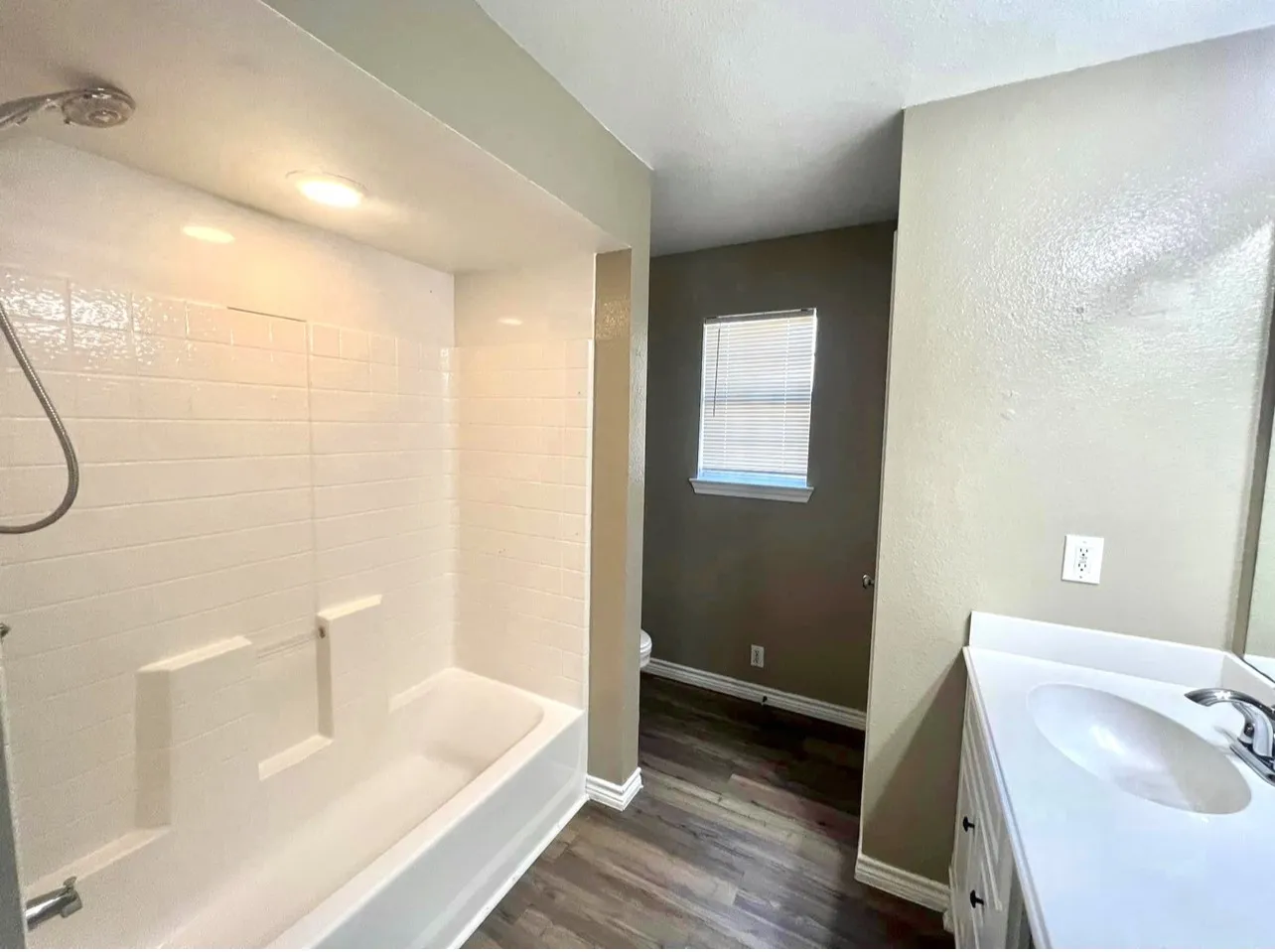Bathroom featuring a white bathtub with subway tile surround, recessed lighting, wood-finish flooring, a white vanity with an integrated sink, and a window with blinds