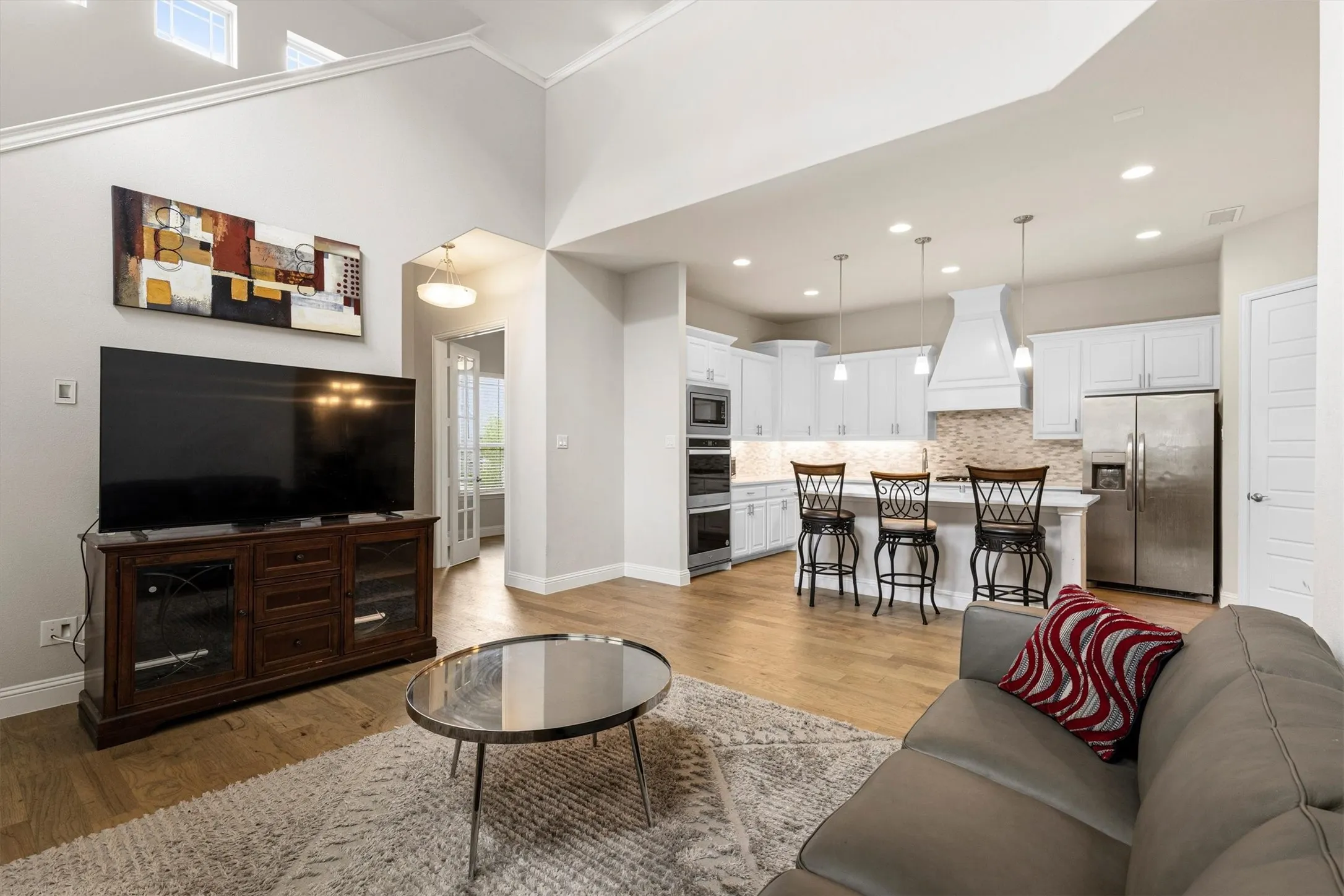 Open-concept living space featuring high vaulted ceilings, light wood-finish flooring, a contemporary kitchen with white cabinetry and stainless steel appliances, and a kitchen island with pendant lighting