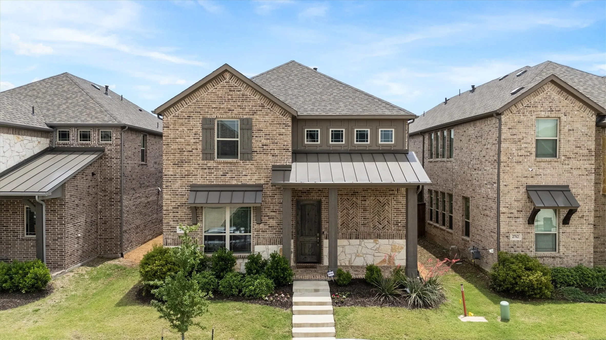 Brick and stone facade residence featuring a covered front porch, grey metal accent awnings, dark wood-finish front door, and architectural shingle roofing