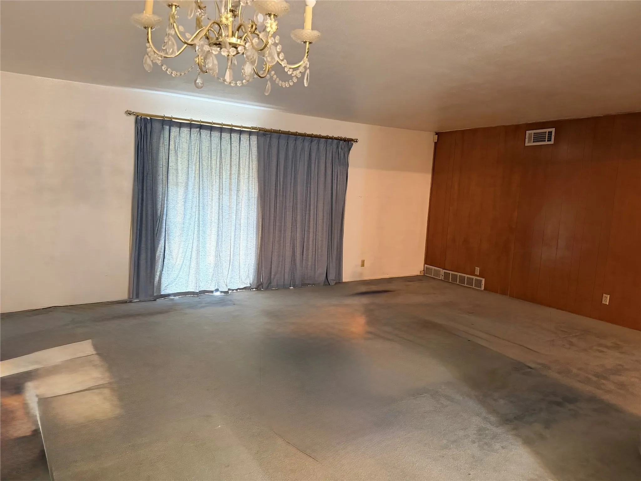 Spacious interior room featuring a decorative chandelier, a large window opening with drapery, and a wall with wood paneling
