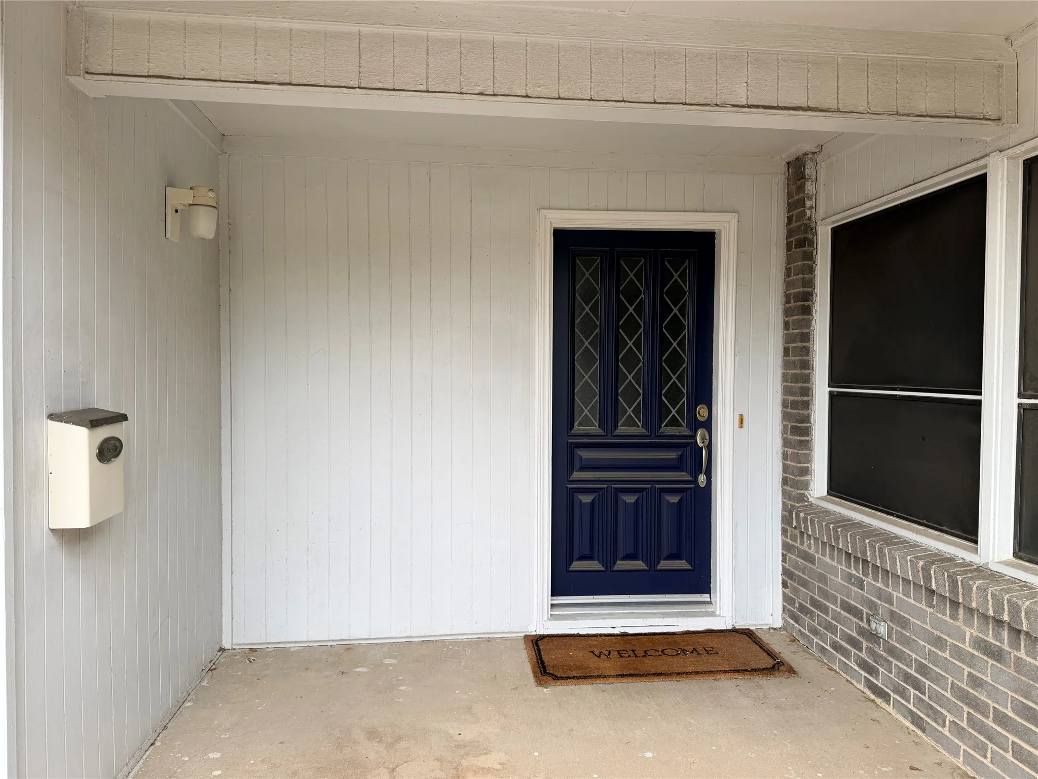 Covered entry featuring a blue paneled door with decorative glass inserts, white vertical siding, and a gray brick facade