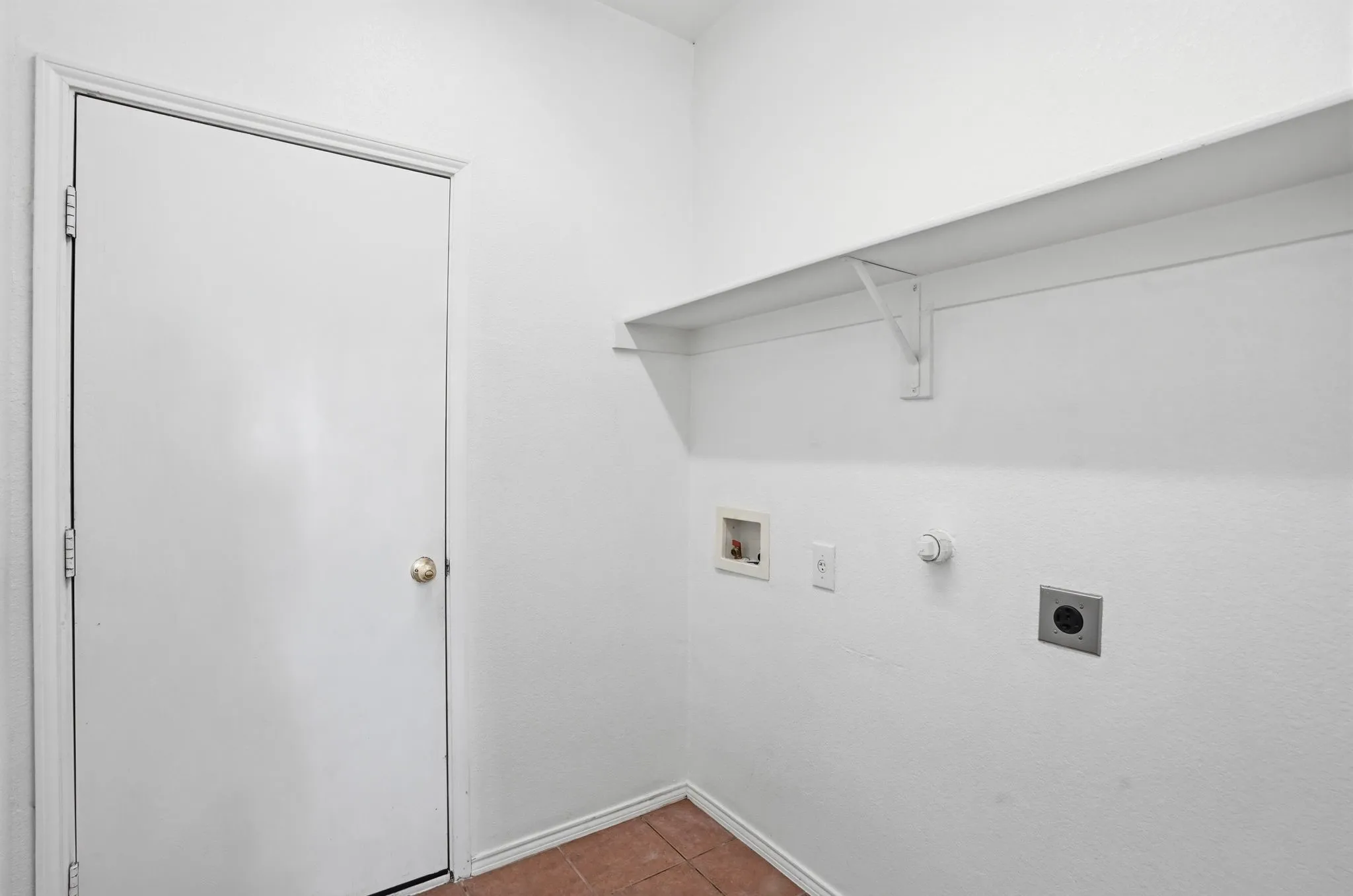 Utility area featuring a white door with a brass knob, a white wall-mounted shelf, and terracotta-toned floor tiling