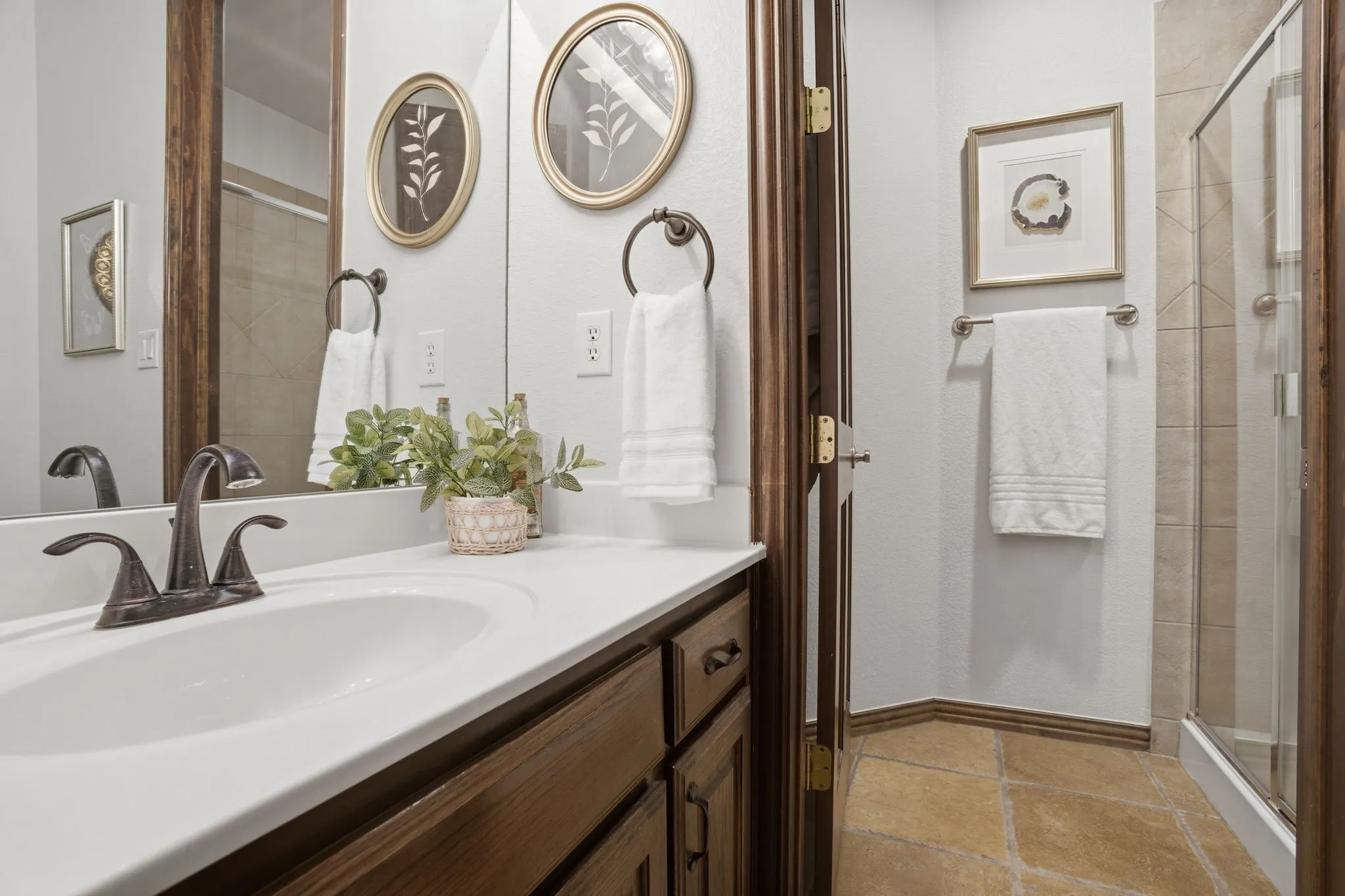 The first-floor full bathroom showcases upgraded travertine flooring and a built-in vanity with warm wood cabinetry, oil rubbed bronze hardware, and a crisp white countertop accented by brushed bronze fixtures. A separate privacy room features a shower with a tile surround, a Pfister faucet and shower head, and a built-in shampoo shelf for added convenience.