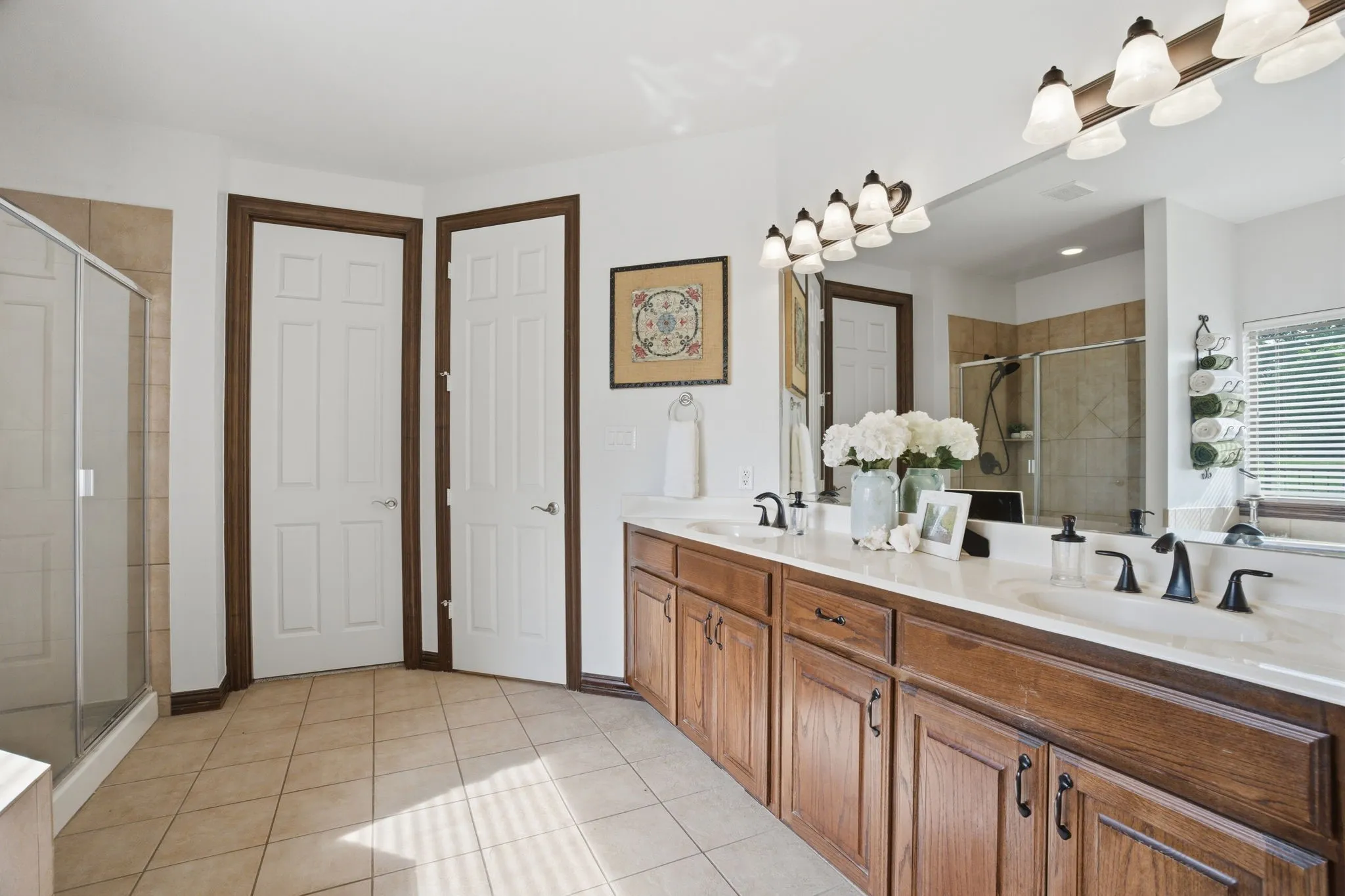 The primary bathroom features tile flooring and a dual sink vanity with warm wood cabinetry, oil rubbed bronze hardware, and a crisp white countertop accented by brushed bronze faucets and fixtures.