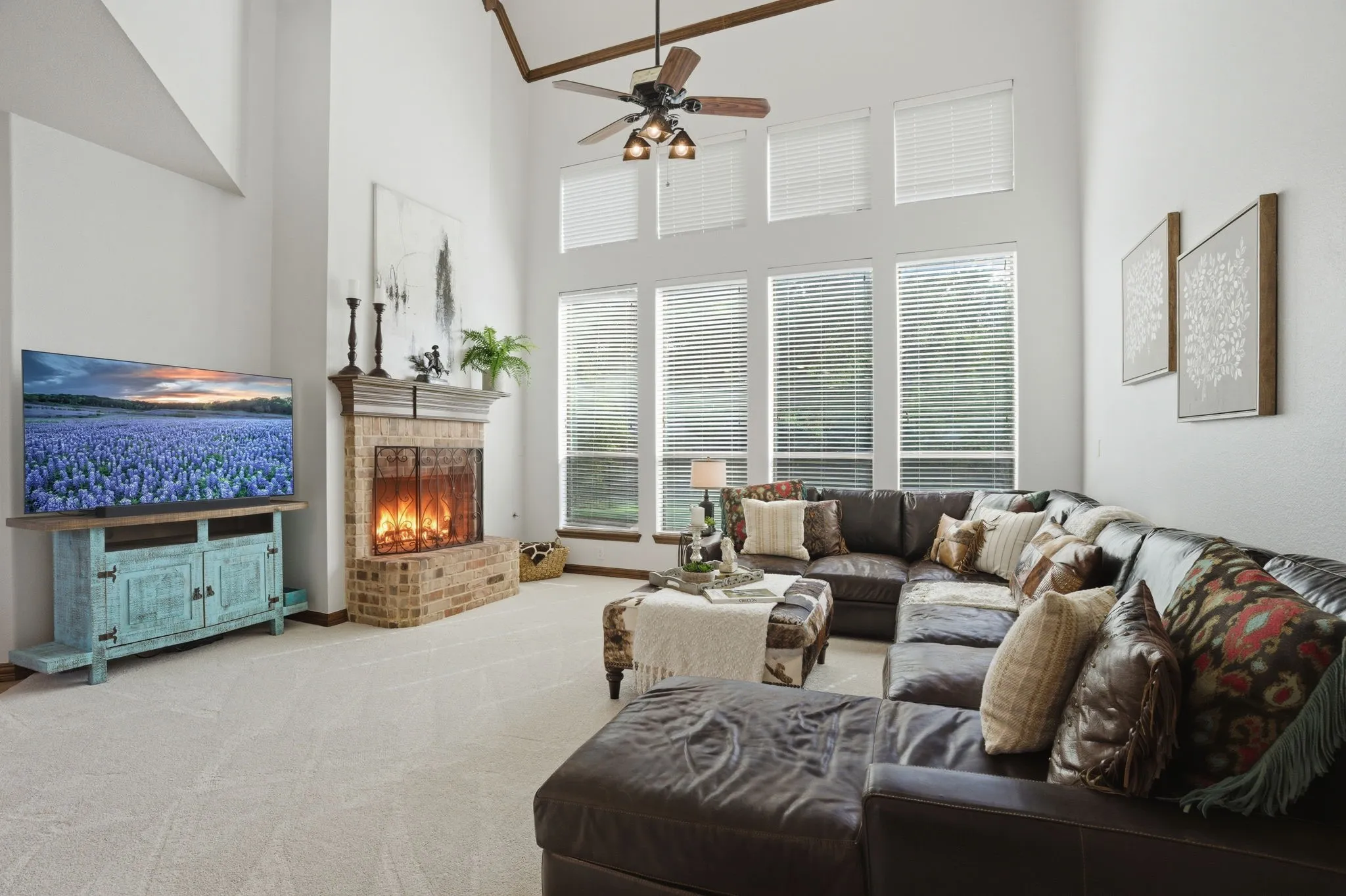The living room offers soaring ceilings, brand new plush carpeting, eight tall windows that frame views of the backyard, and a brick gas fireplace with a raised hearth and wood mantel as a warm focal point. The vaulted ceiling with crown molding and a ceiling fan enhances the room’s sense of space and comfort.

