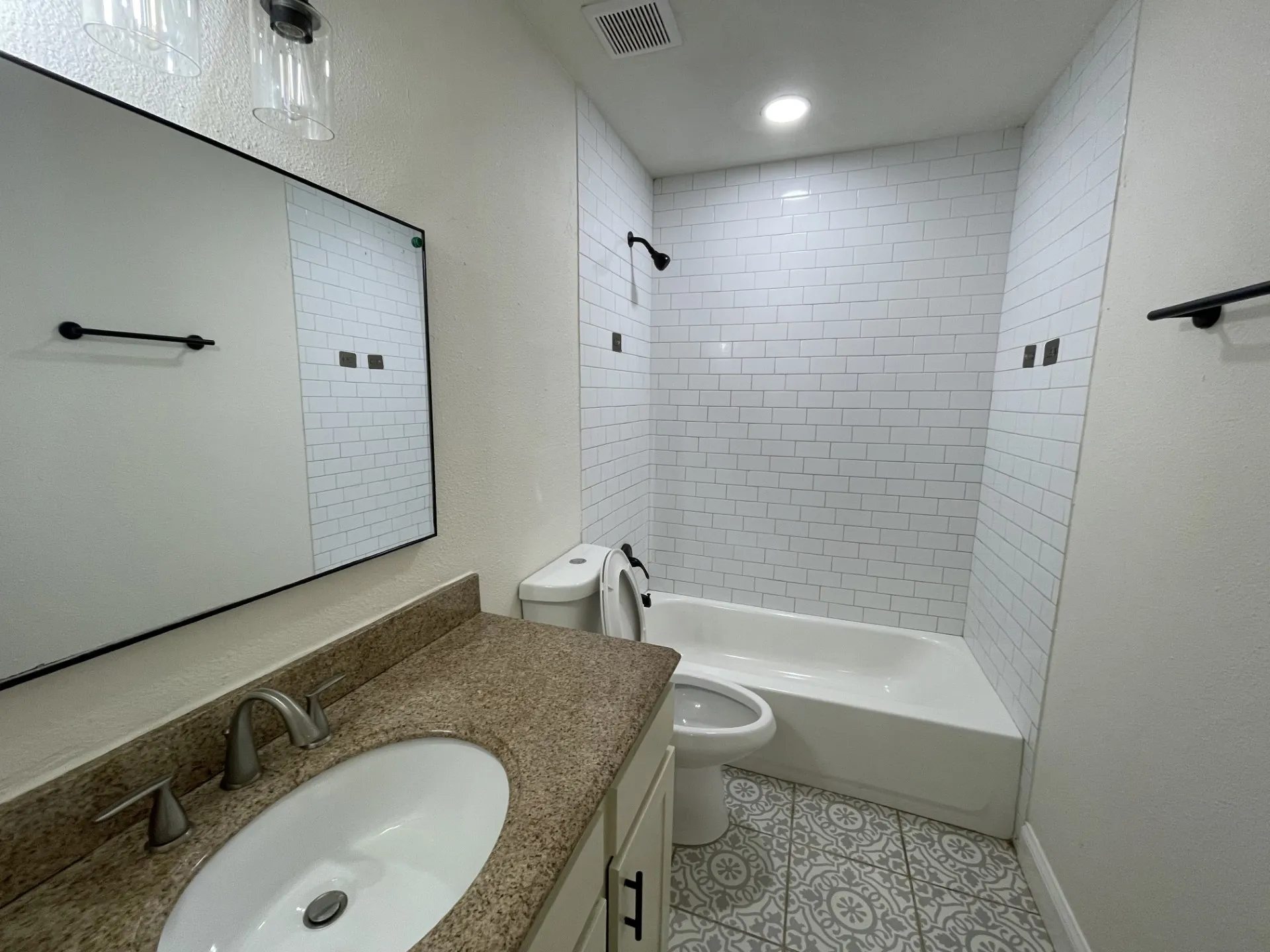 Bathroom featuring a white subway tile shower surround, patterned floor tiles, a vanity with a stone-finish countertop, a single basin sink, and a large wall mirror