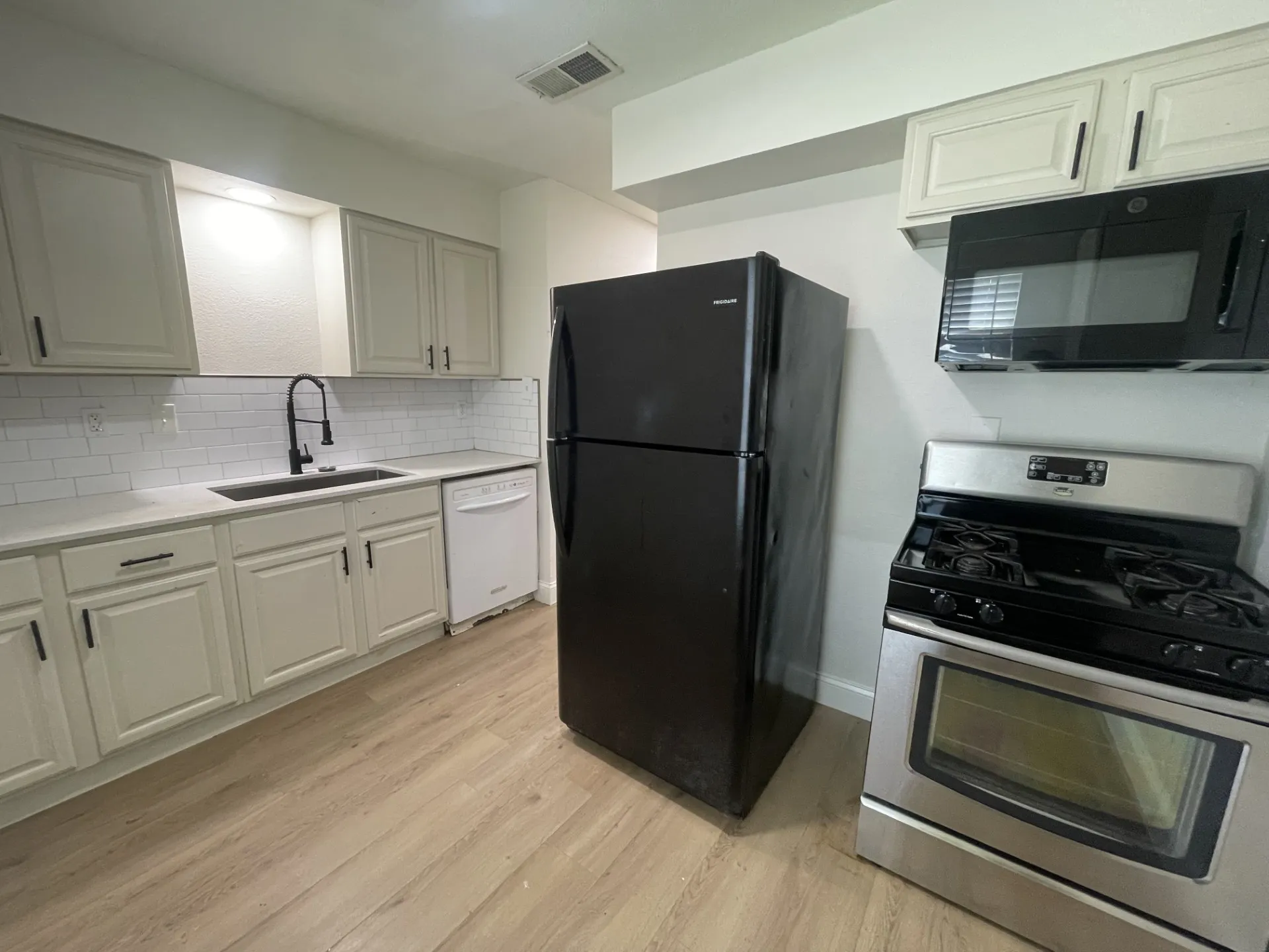 Kitchen featuring wood-finish flooring, white subway tile backsplash, light-colored cabinetry with dark hardware, a black refrigerator, and a stainless steel range