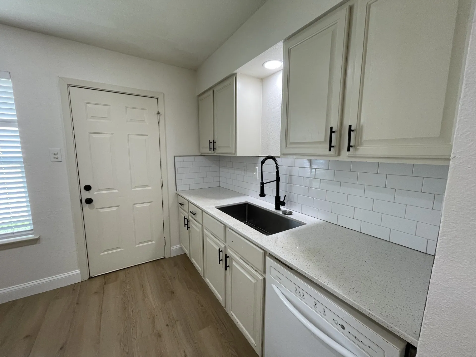 Kitchen featuring wood-finish flooring, white cabinetry with black hardware, white subway tile backsplash, light speckled countertops, and a black gooseneck faucet