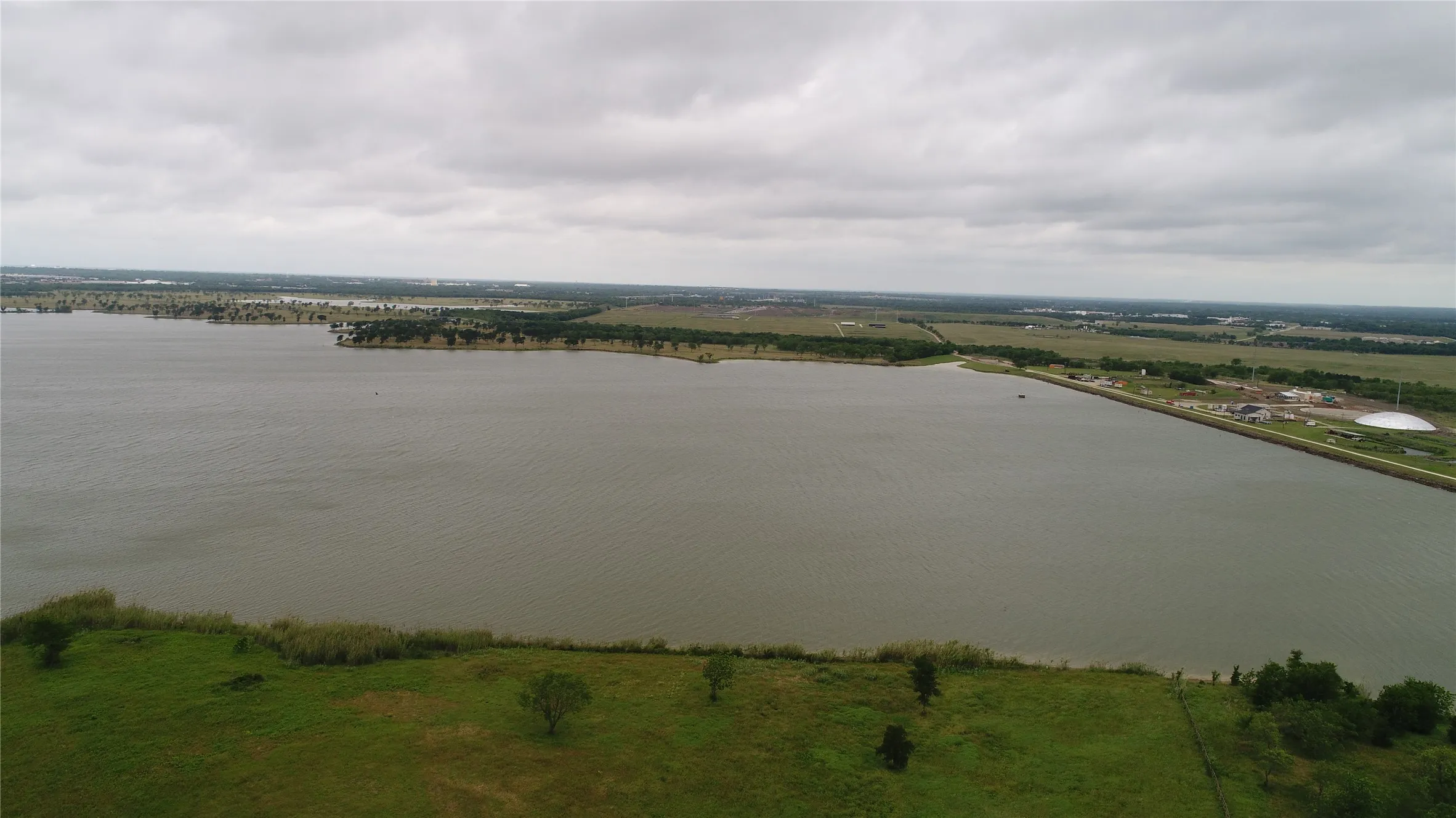 Expansive water body with surrounding green land and distant structures under a cloudy sky