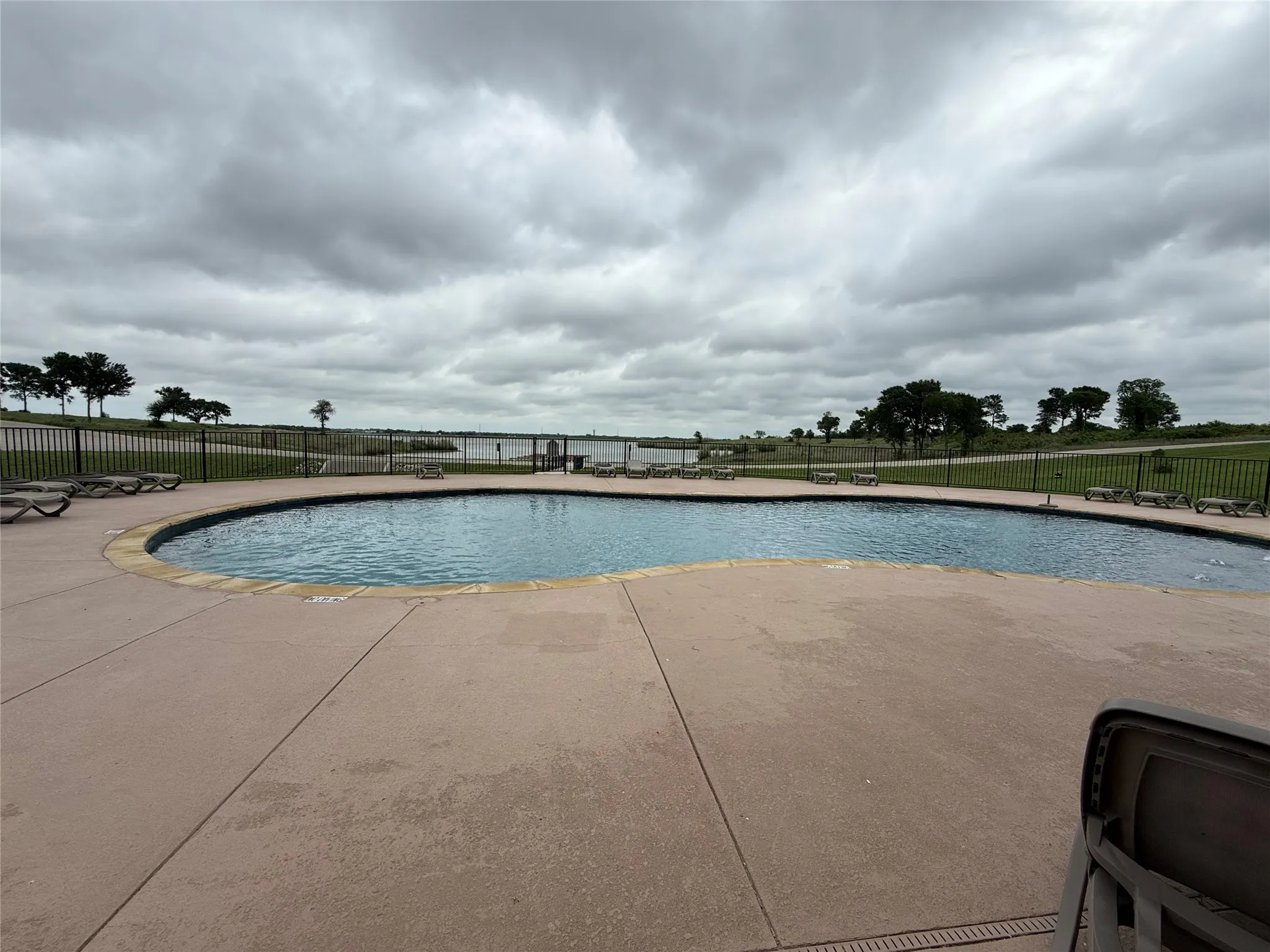 Freeform swimming pool with concrete decking, surrounded by a black metal fence with open view to the lake.