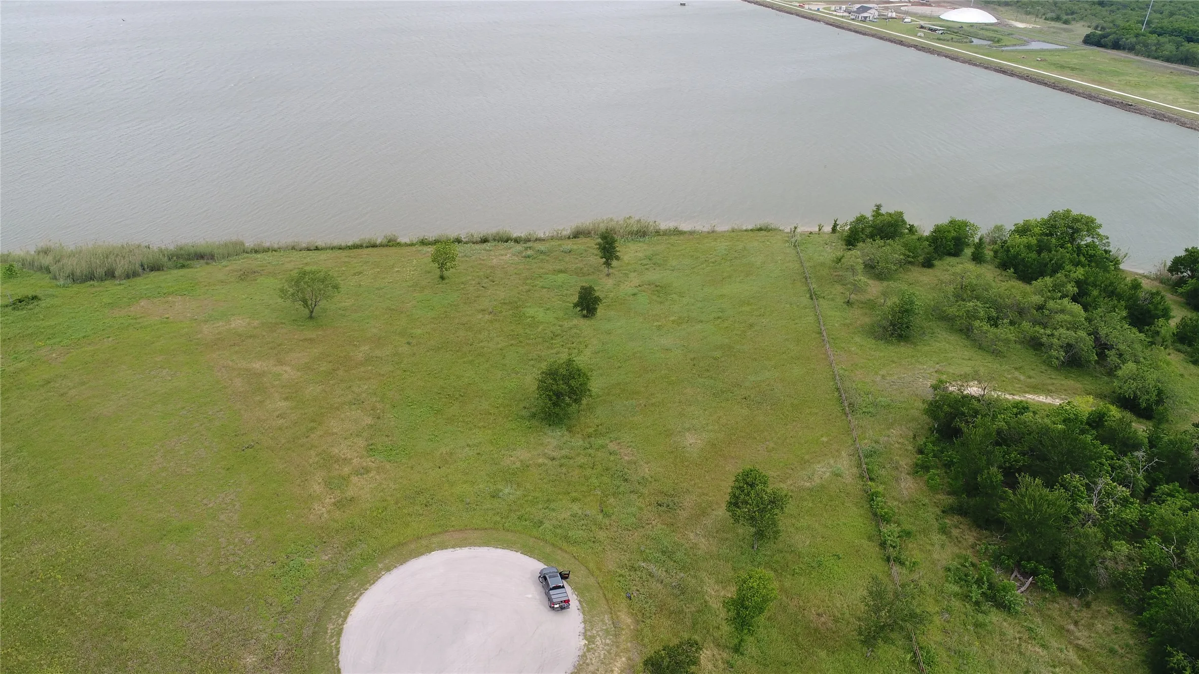 Expansive waterfront lot featuring a large body of water, green landscape, scattered trees, and a circular gravel drive